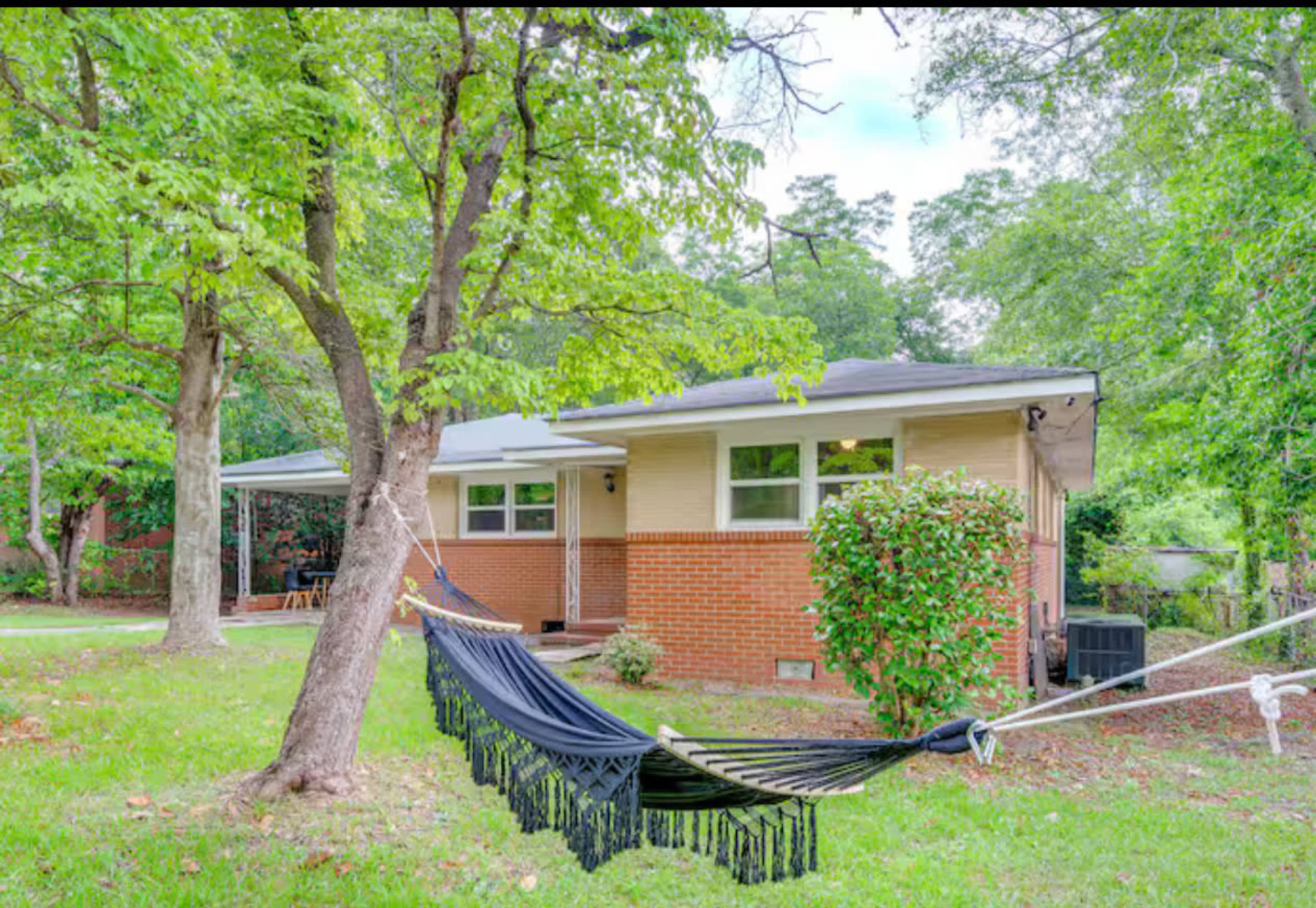 A hammock is strung between two trees in a grassy yard beside a brick house.