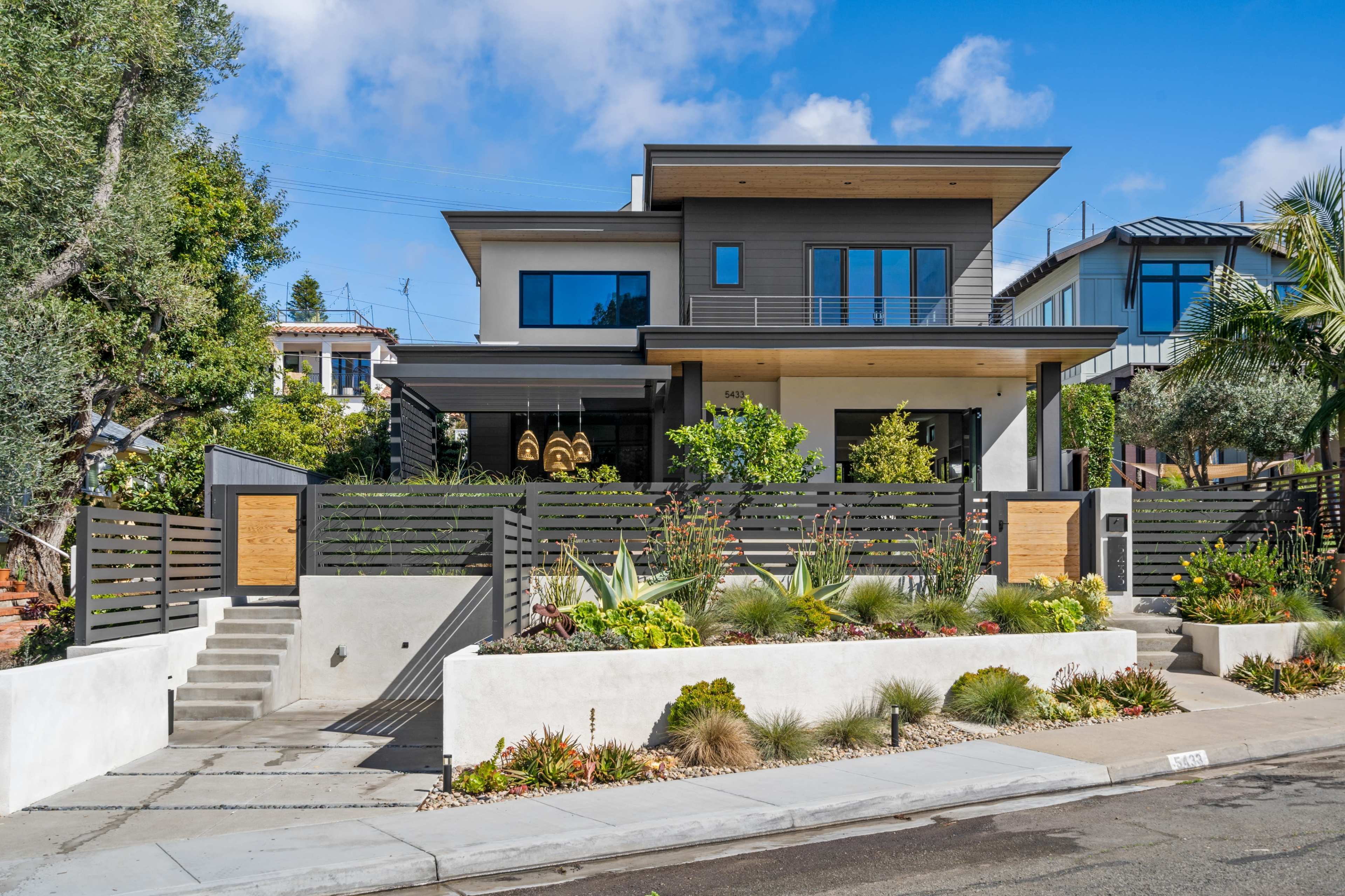 A modern two-story house with a landscaped front yard, featuring a combination of plants and stone walkways, bordered by a wooden fence.