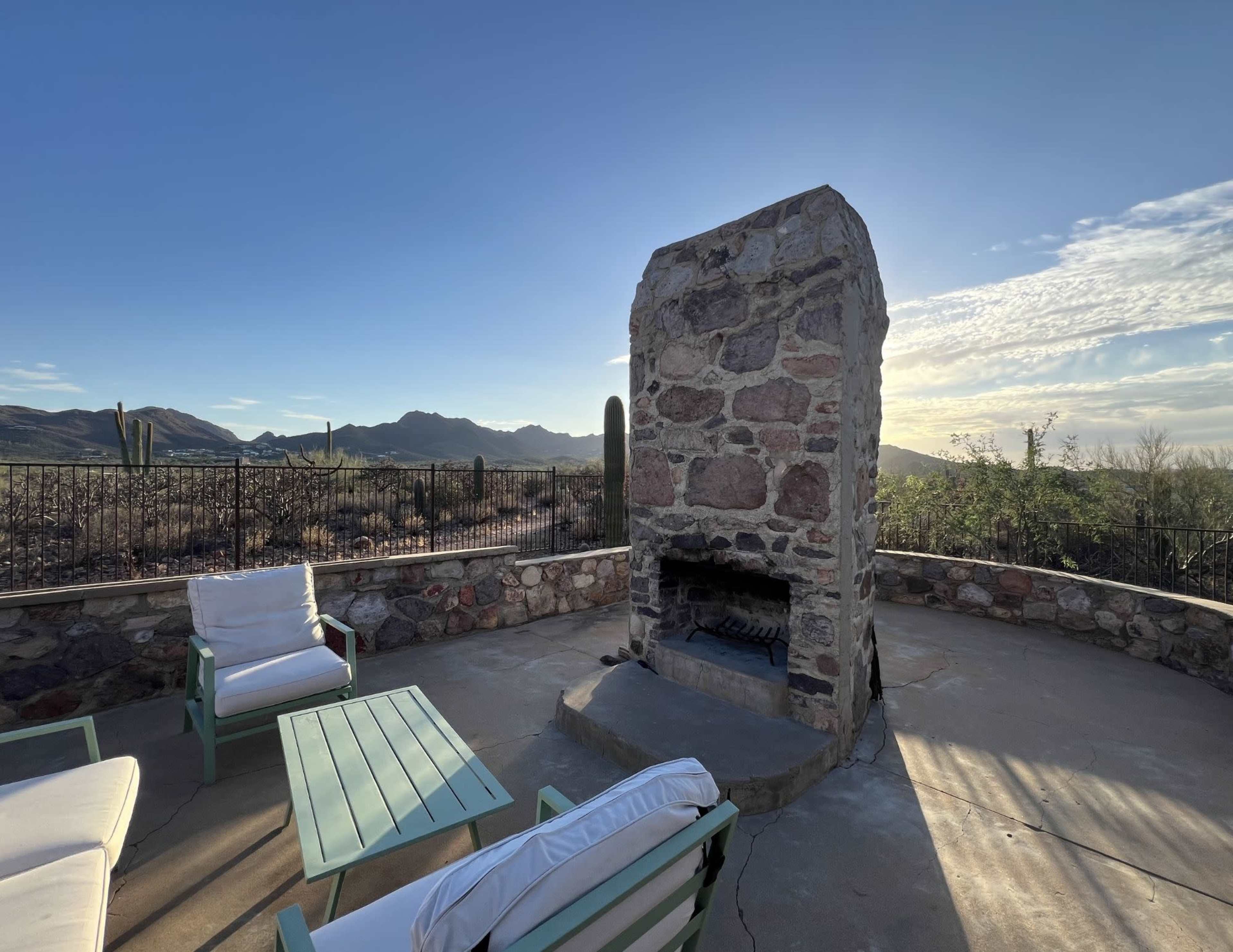 The image shows a stone outdoor fireplace surrounded by seating on a patio, with a mountainous desert landscape in the background.