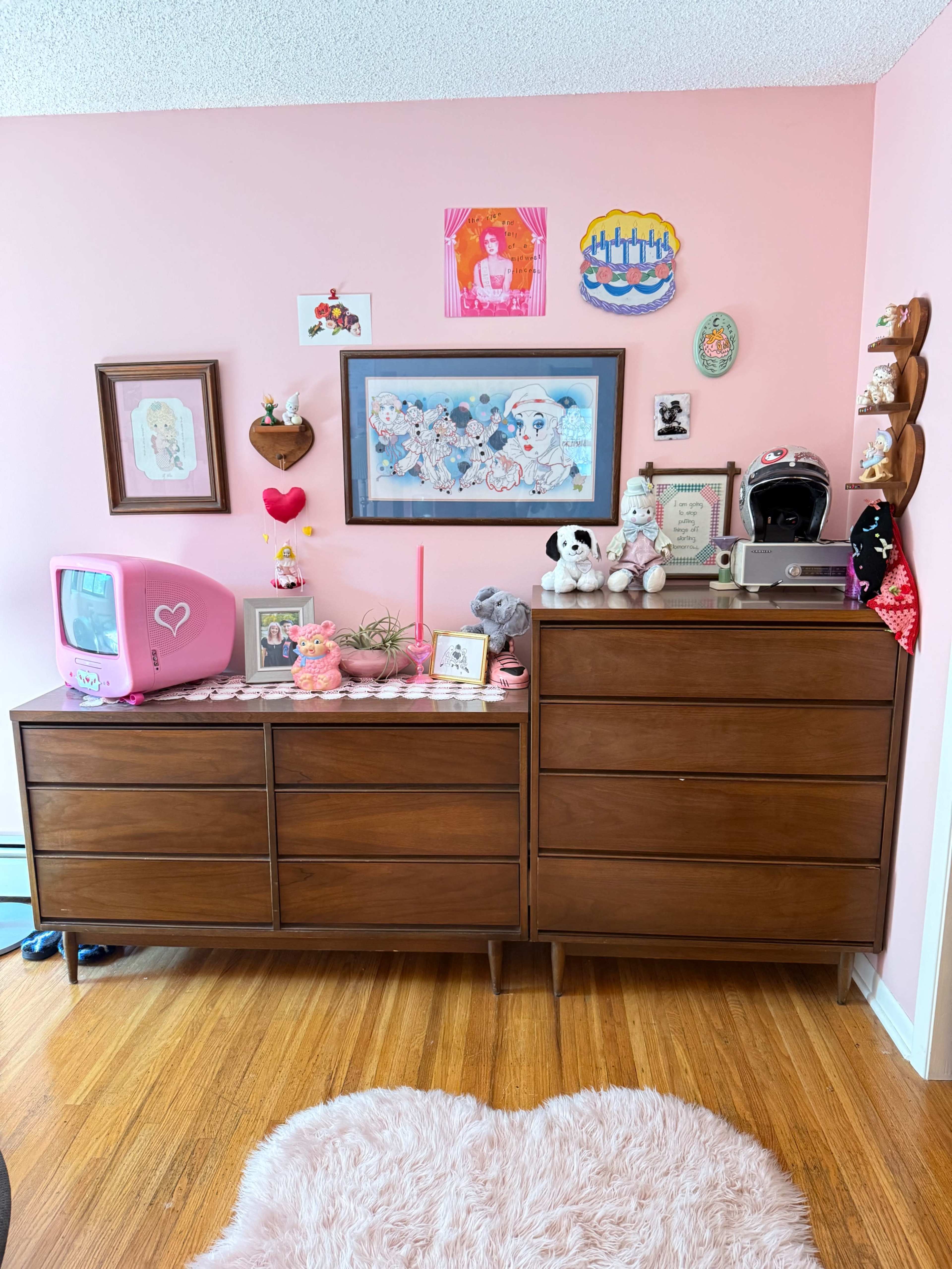 A wooden dresser is adorned with various decorative items and framed art against a pink wall.