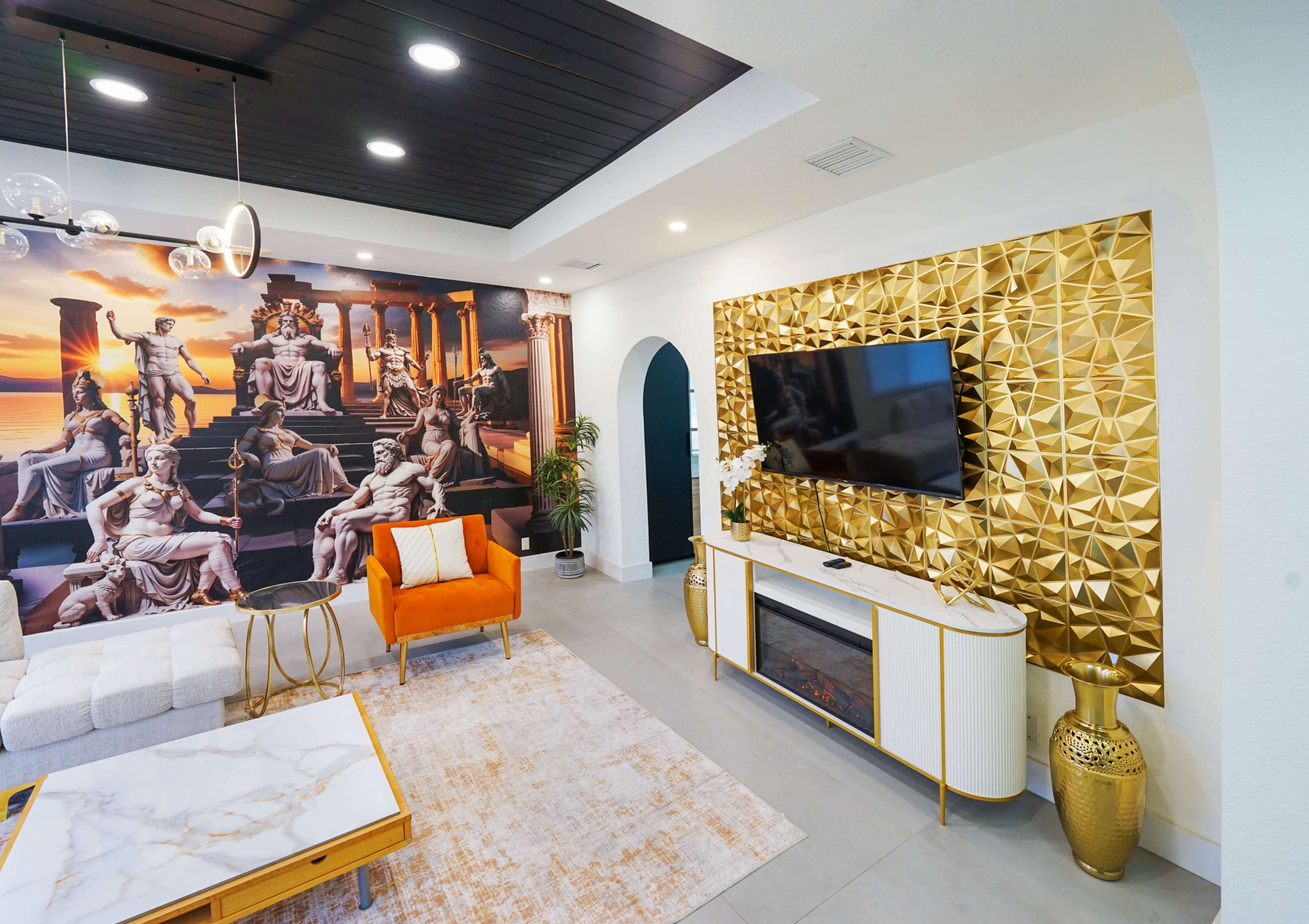 The image shows a modern living room with a large mural featuring classical sculptures, a gold-accented television console, and an orange armchair.