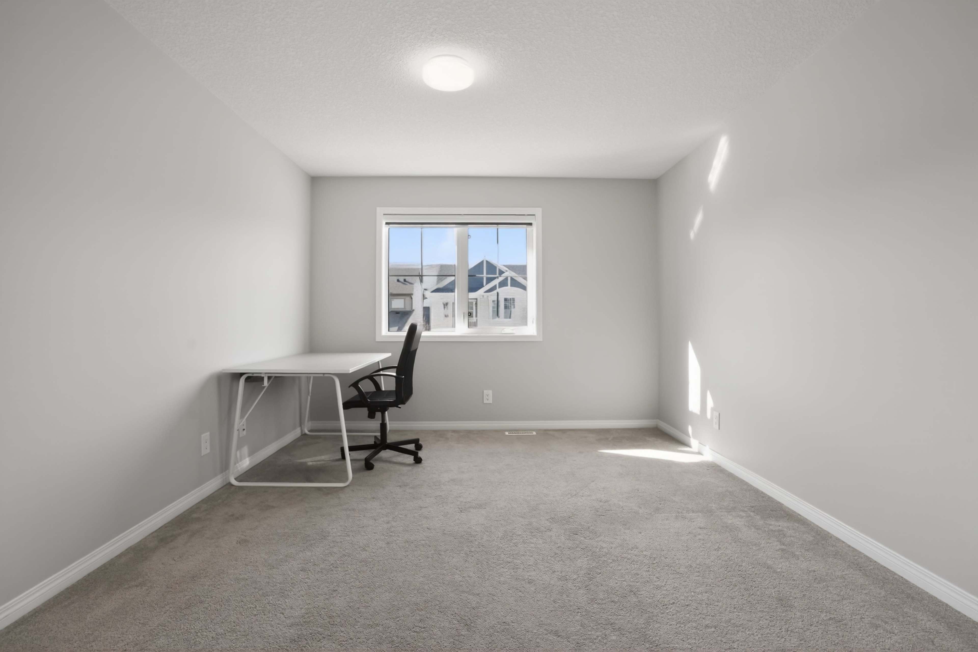 The image shows a sparsely furnished room with a desk and chair next to a window, featuring light grey walls and carpet.