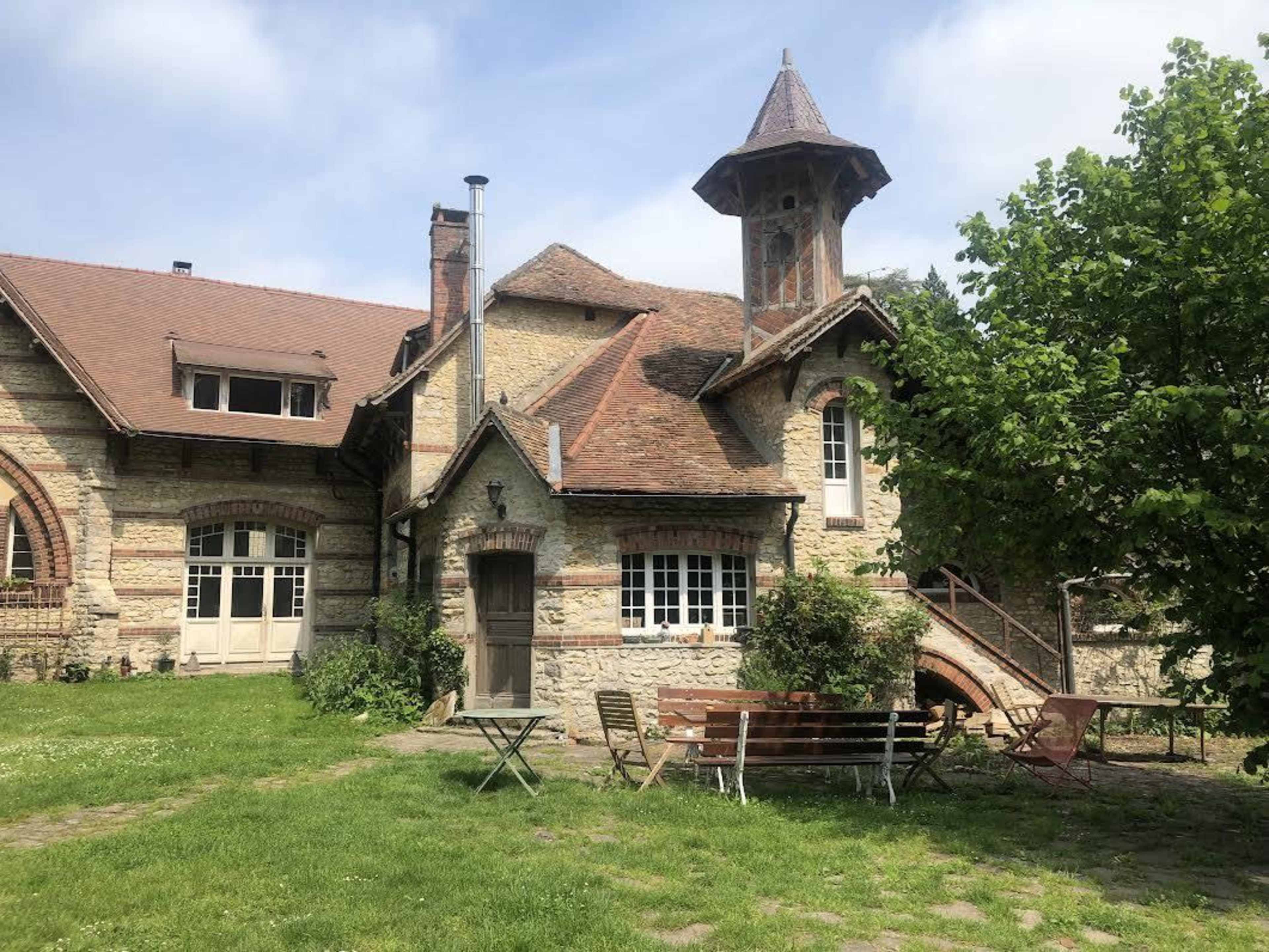 The image shows a historic building with a turret, surrounded by green grass and trees, featuring an outdoor seating area with chairs and a table.