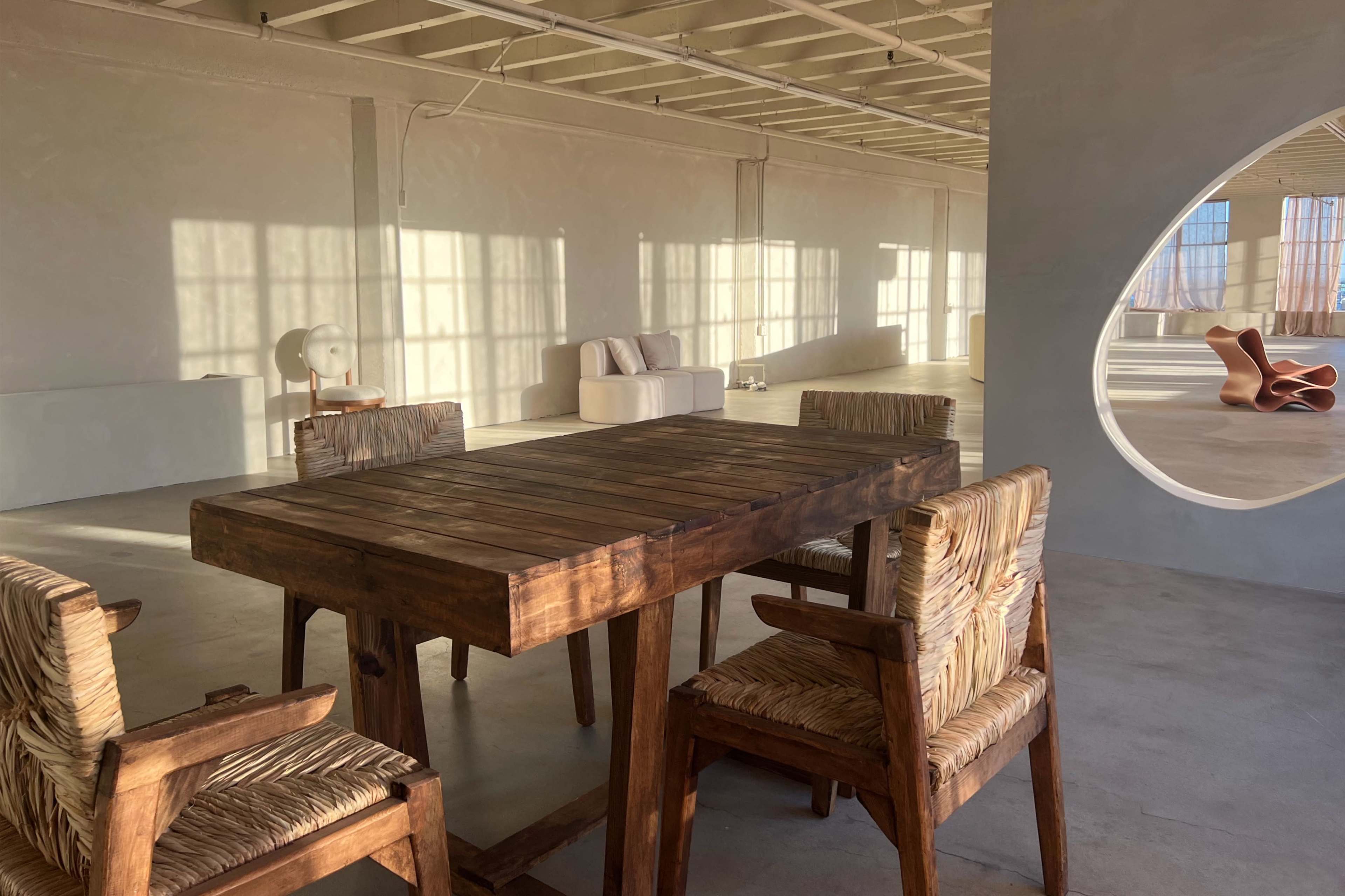 The image shows a minimalist interior space featuring a wooden dining table surrounded by four chairs with woven seats, alongside large windows casting shadows on the floor.