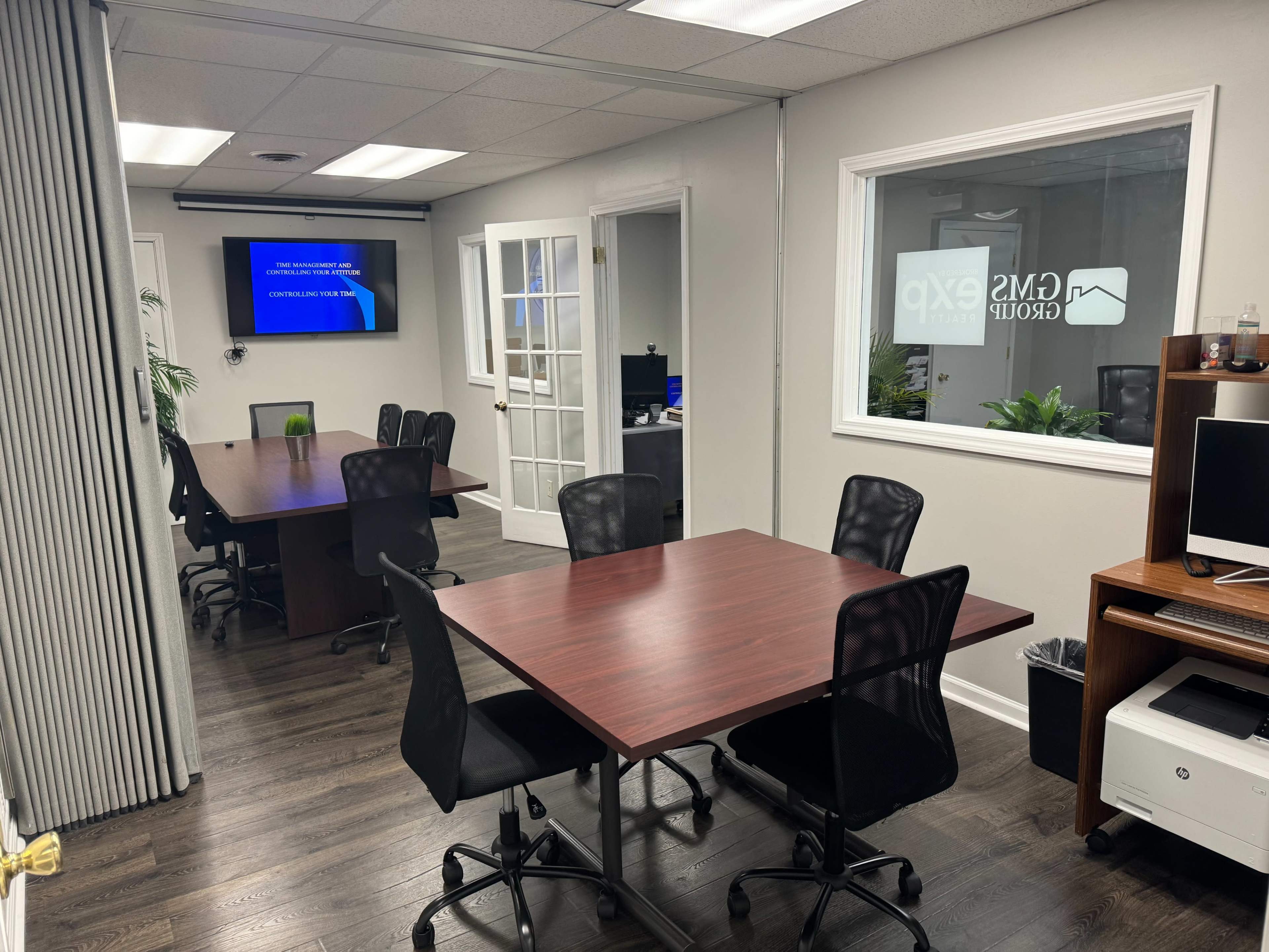 A modern office space with multiple desks arranged around a central area, featuring a presentation screen and large windows.