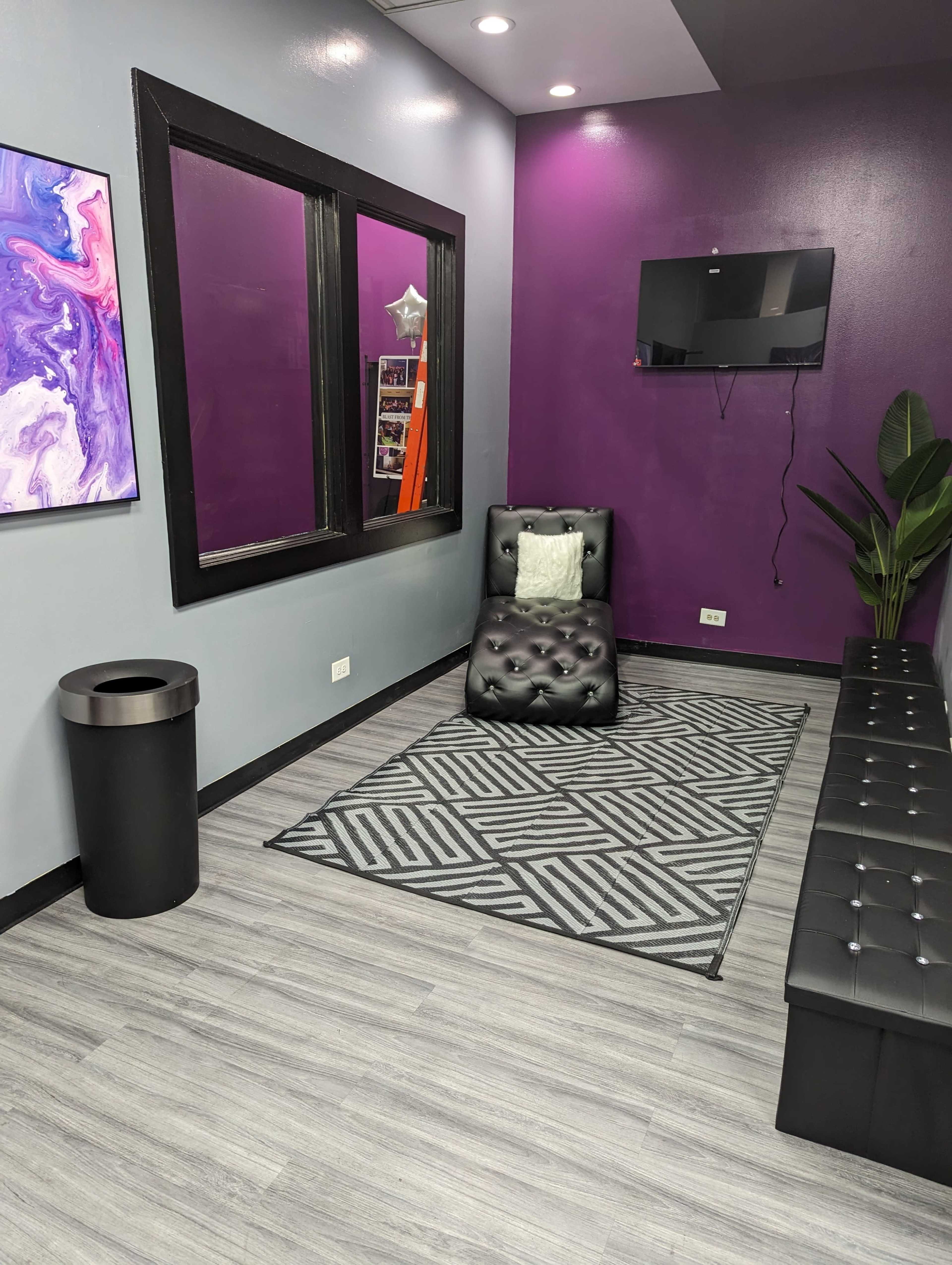 A small waiting area with a black leather chair, a gray patterned rug, a trash can, a wall-mounted TV, and a purple accent wall.