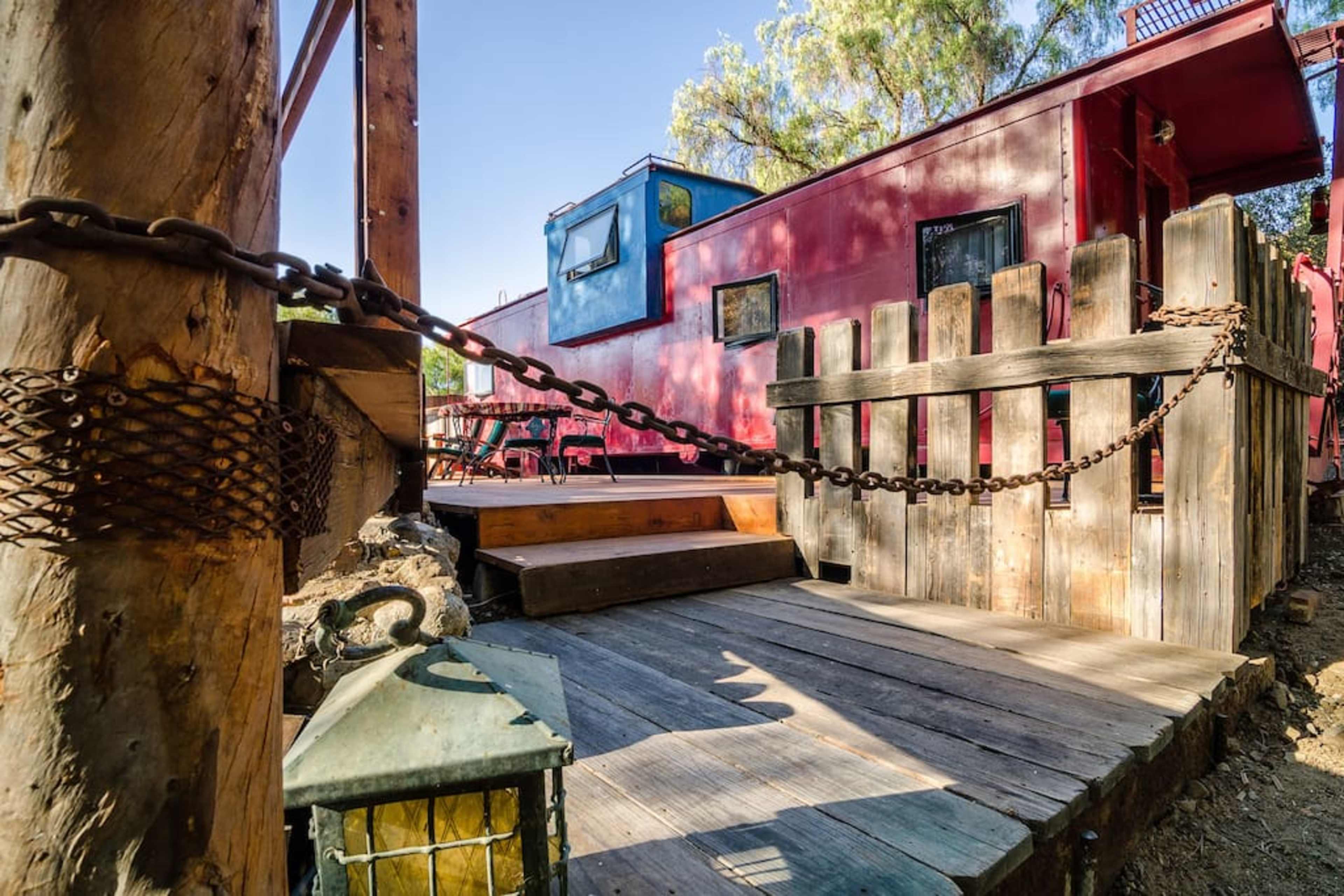 A red train caboose beside a wooden deck with a chain-link fence and a lantern on the ground.