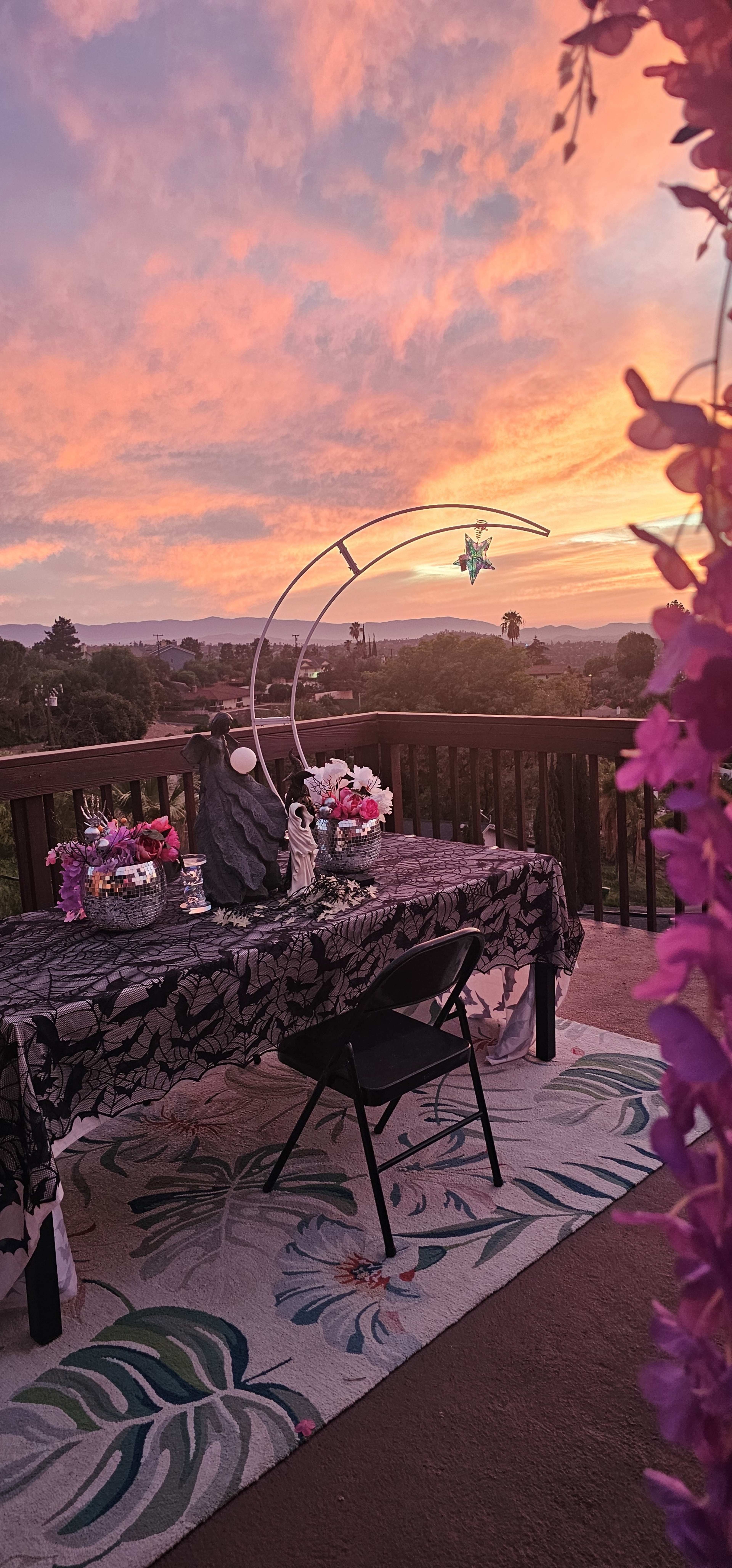 A table with floral decorations is set against a colorful sunset sky, with a view of trees and distant hills in the background.