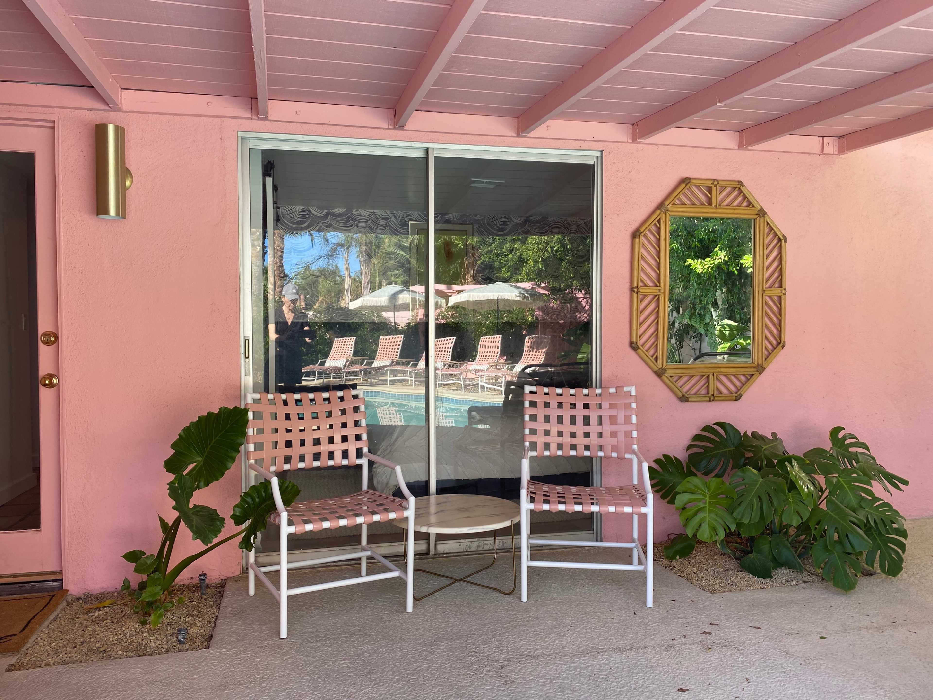 Two white chairs with pink accents and a small table are positioned outside a glass door that reflects a swimming pool area with loungers under umbrellas.