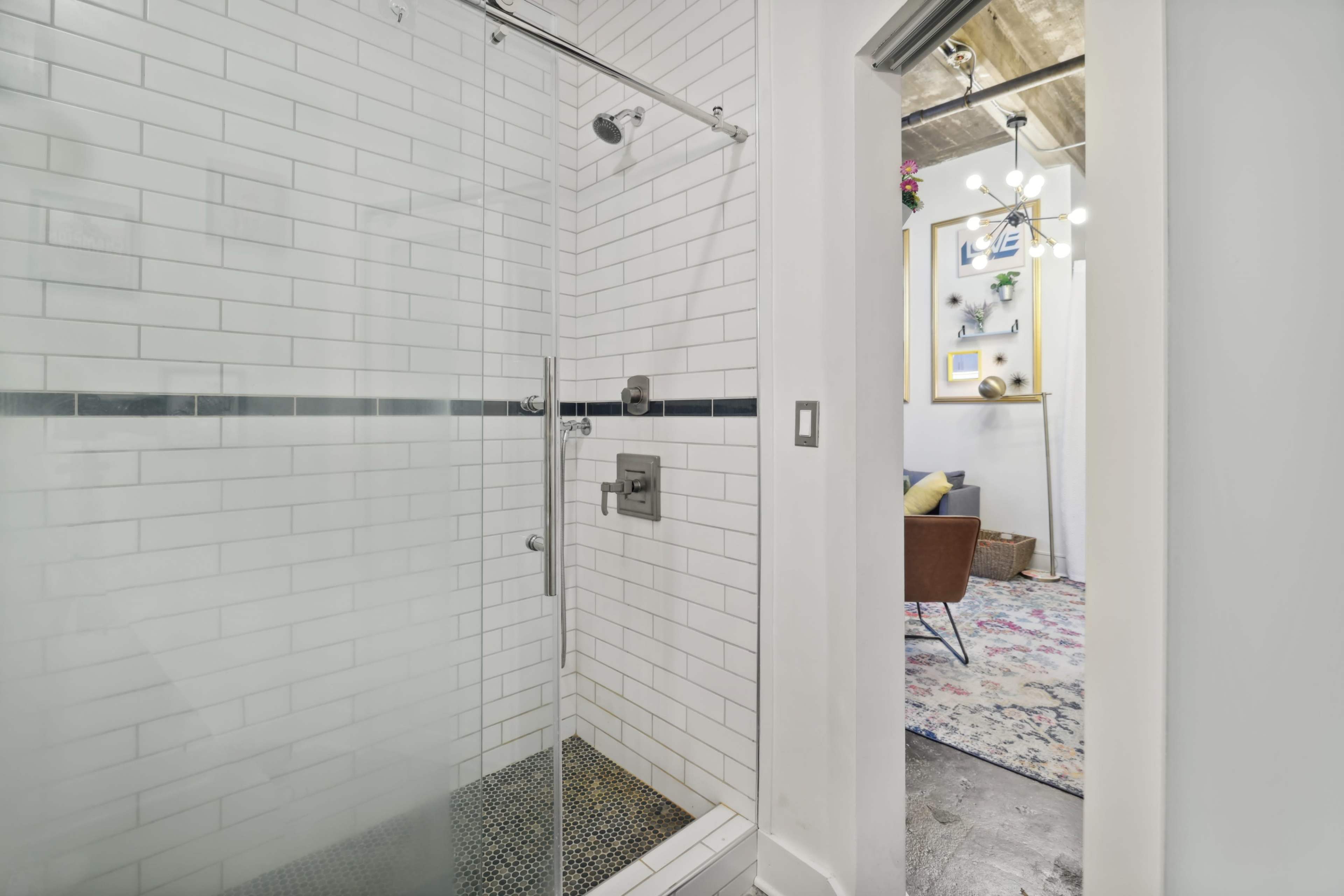 A glass-enclosed shower with white subway tiles and a dark tile floor, adjacent to a room with a colorful rug and modern furniture visible in the background.