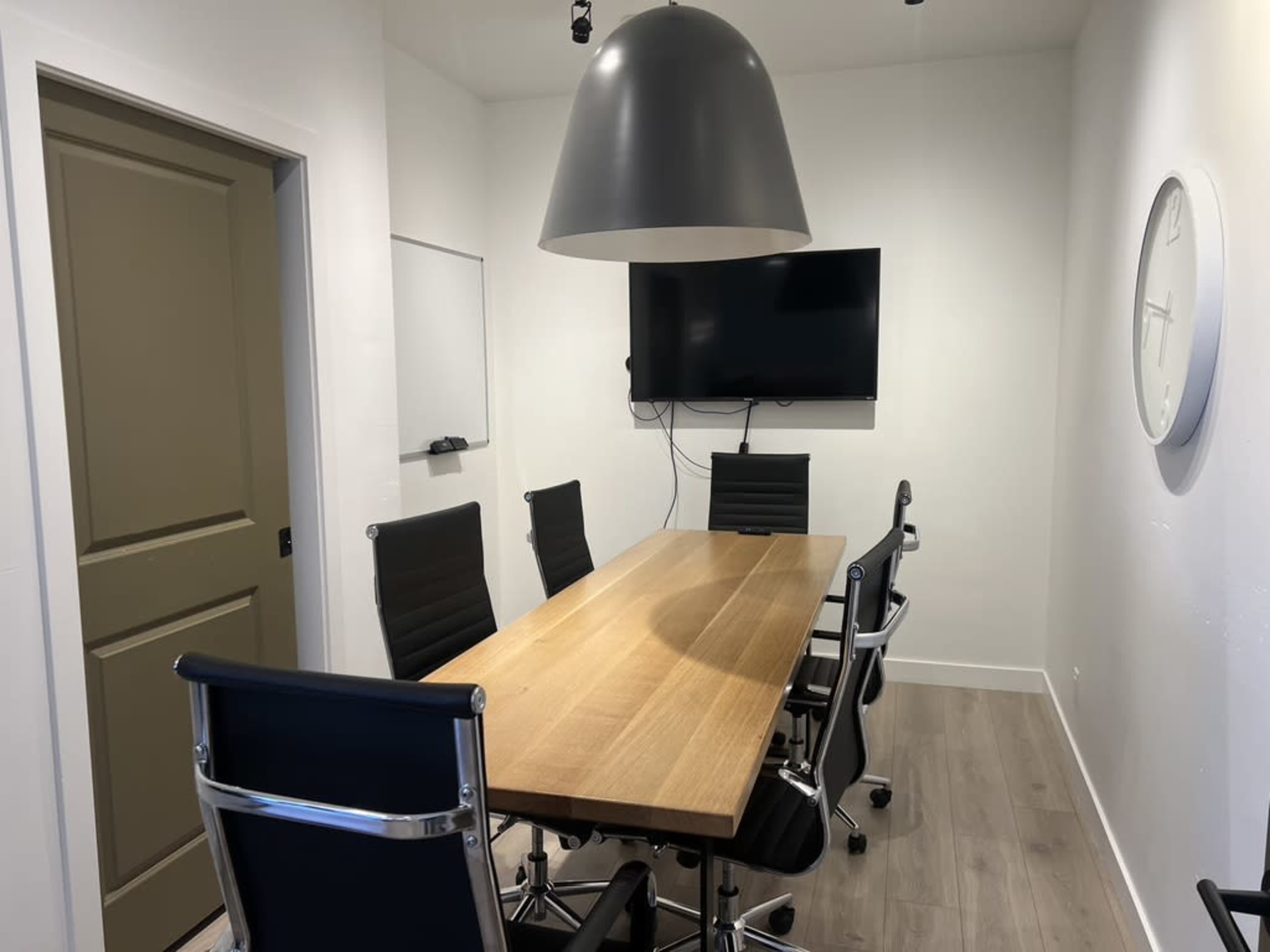 A modern meeting room features a wooden conference table surrounded by black ergonomic chairs, a wall-mounted TV, a whiteboard, and a large pendant light.