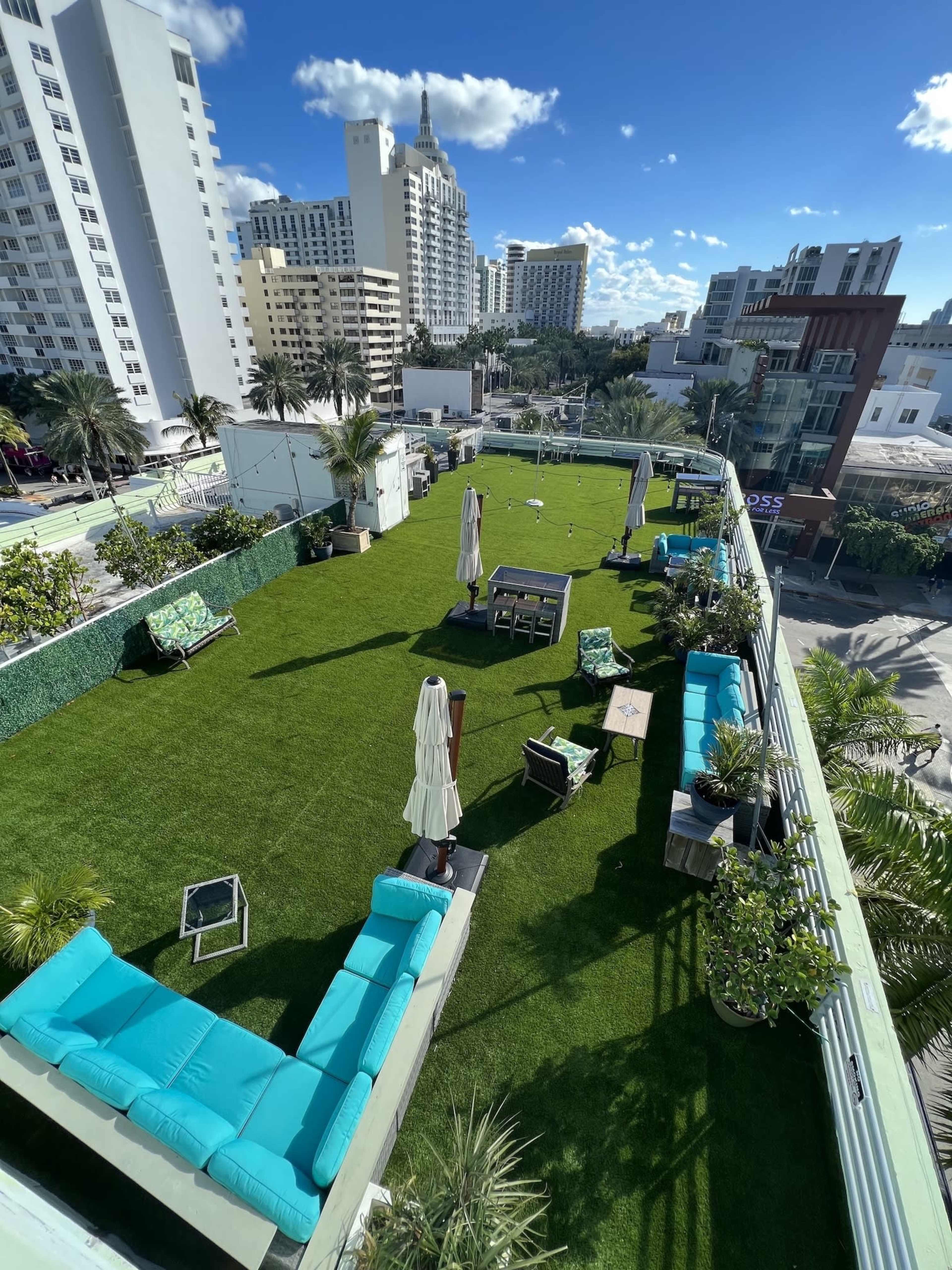 The image shows a rooftop garden with green artificial grass, seating areas, and palm trees, overlooking a cityscape of palm-lined streets and buildings.