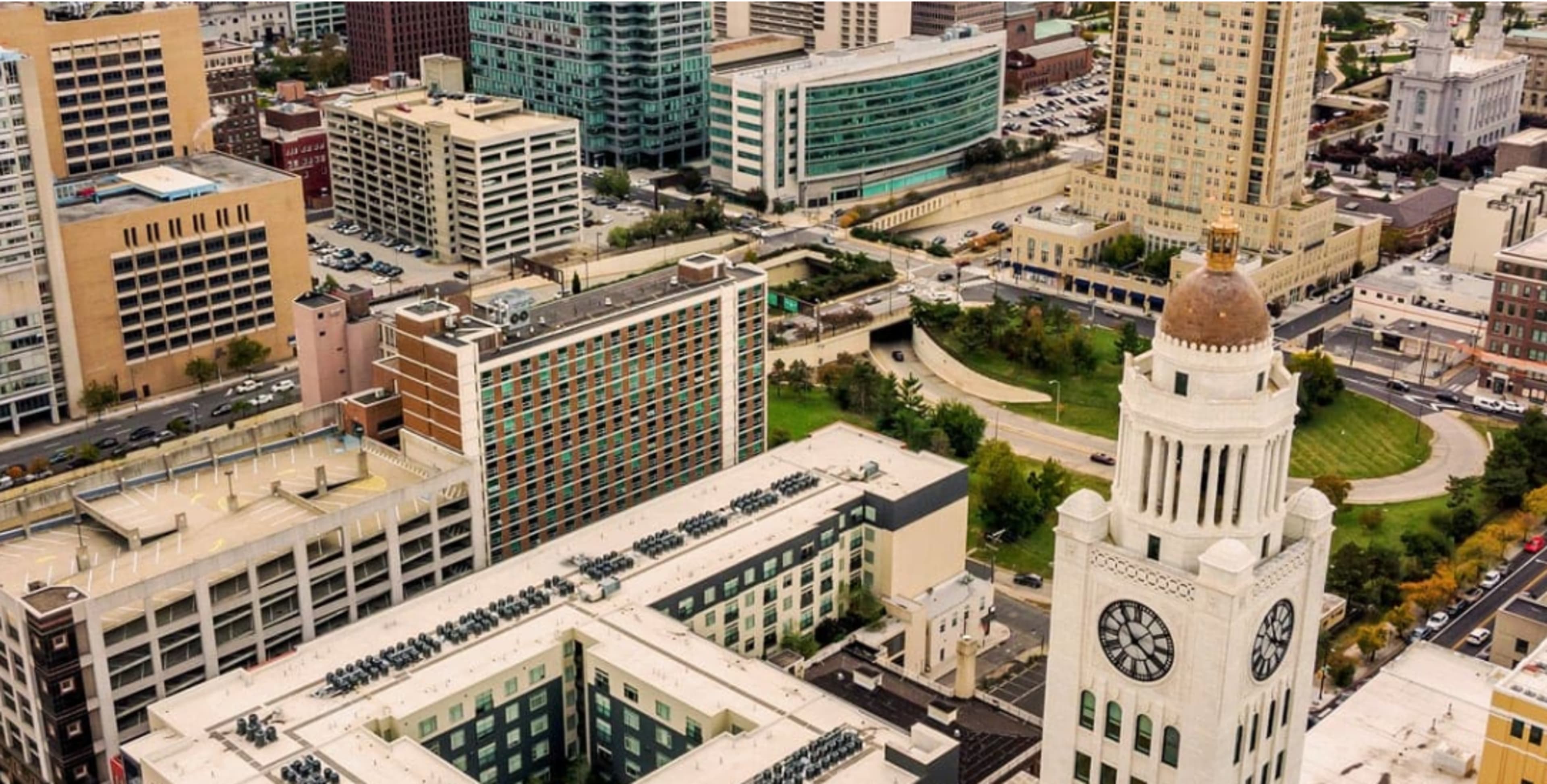 The image shows a view of a cityscape featuring a mix of modern buildings and a prominent clock tower surrounded by green space.