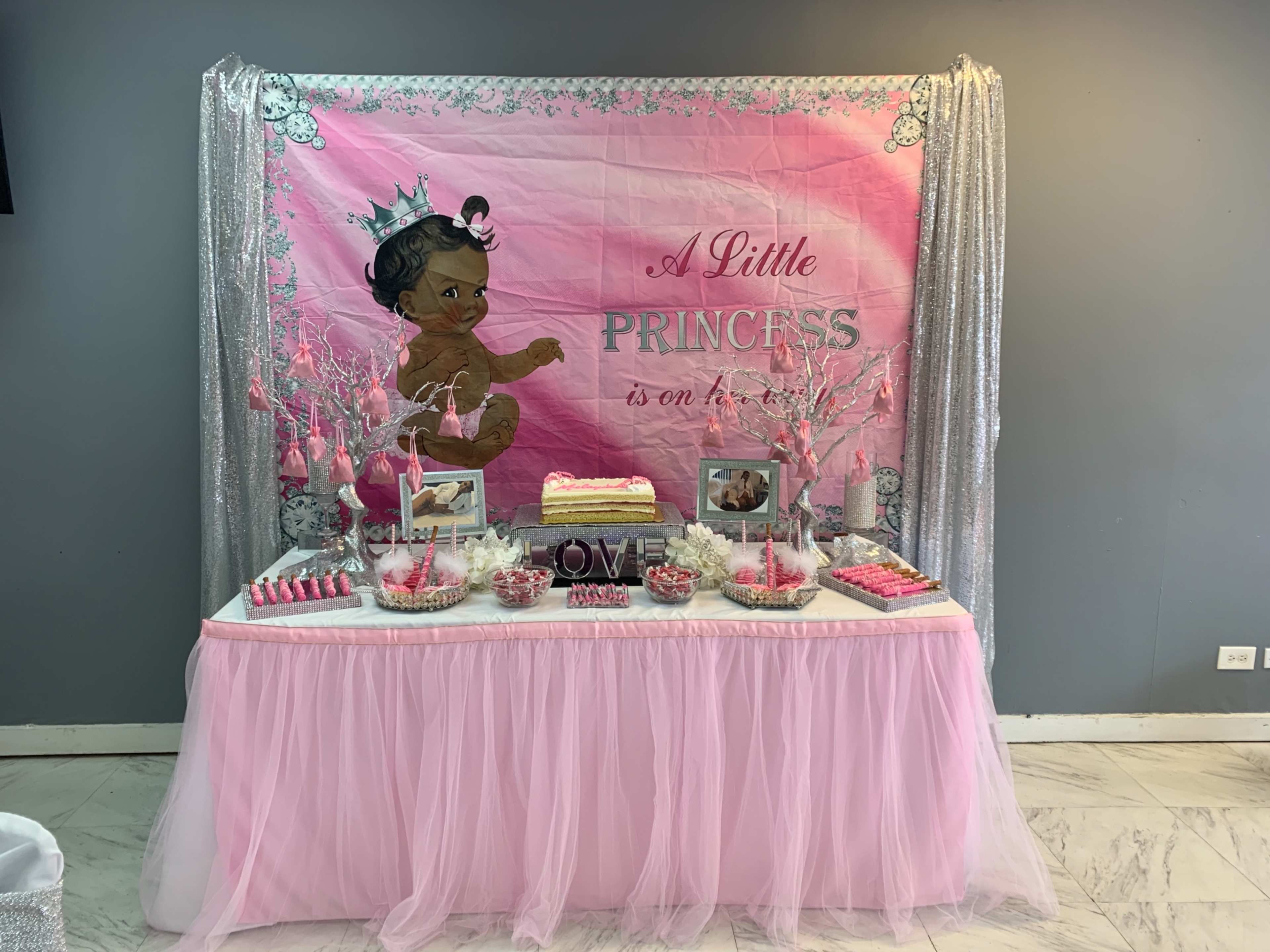 A decorated table featuring a pink-themed birthday backdrop with a cake and various sweet treats arranged on white and silver table covers.