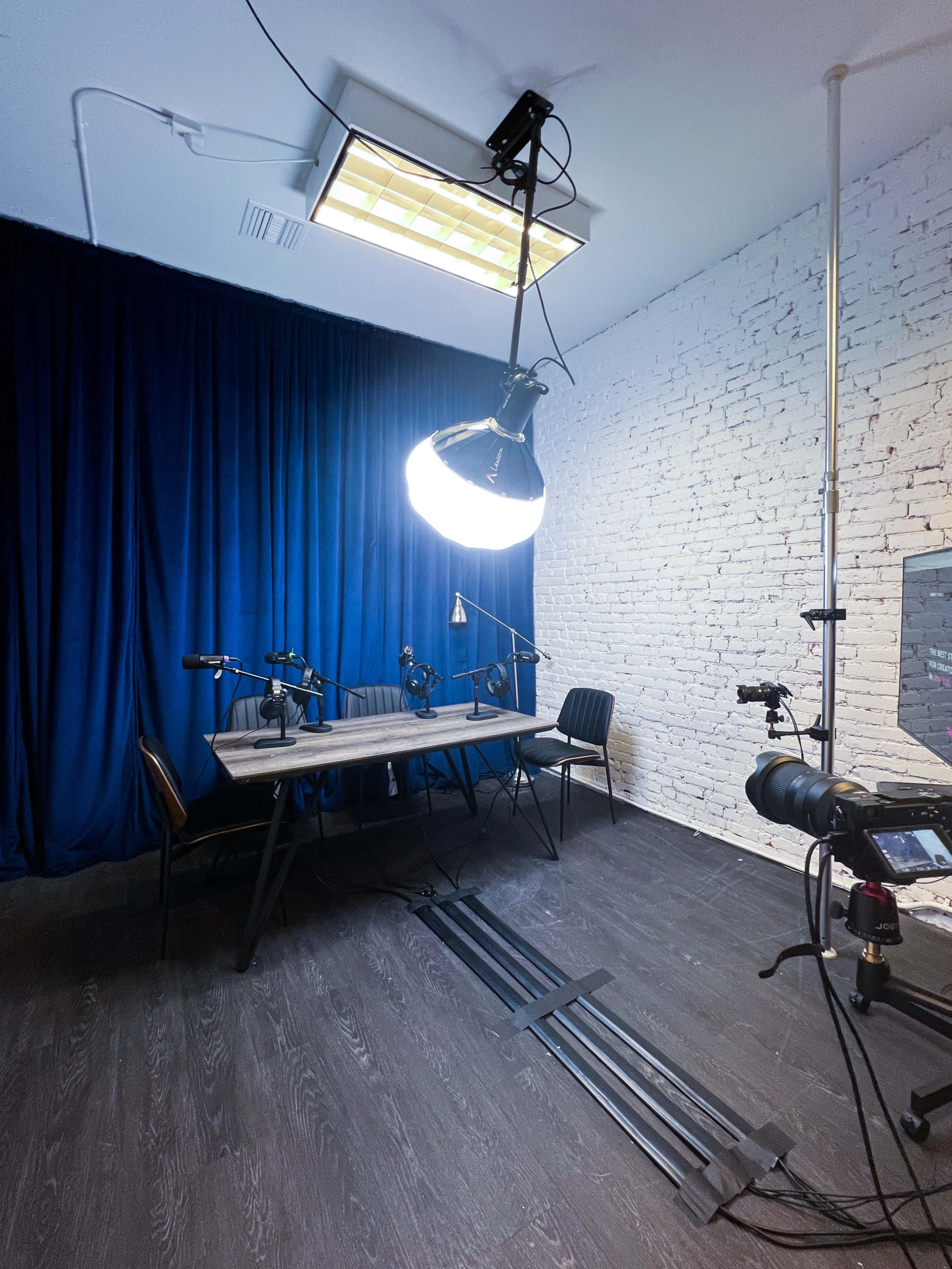 A dimly lit recording studio features a large circular light above a table with multiple microphones and a chair, set against a backdrop of blue curtains and a white brick wall.