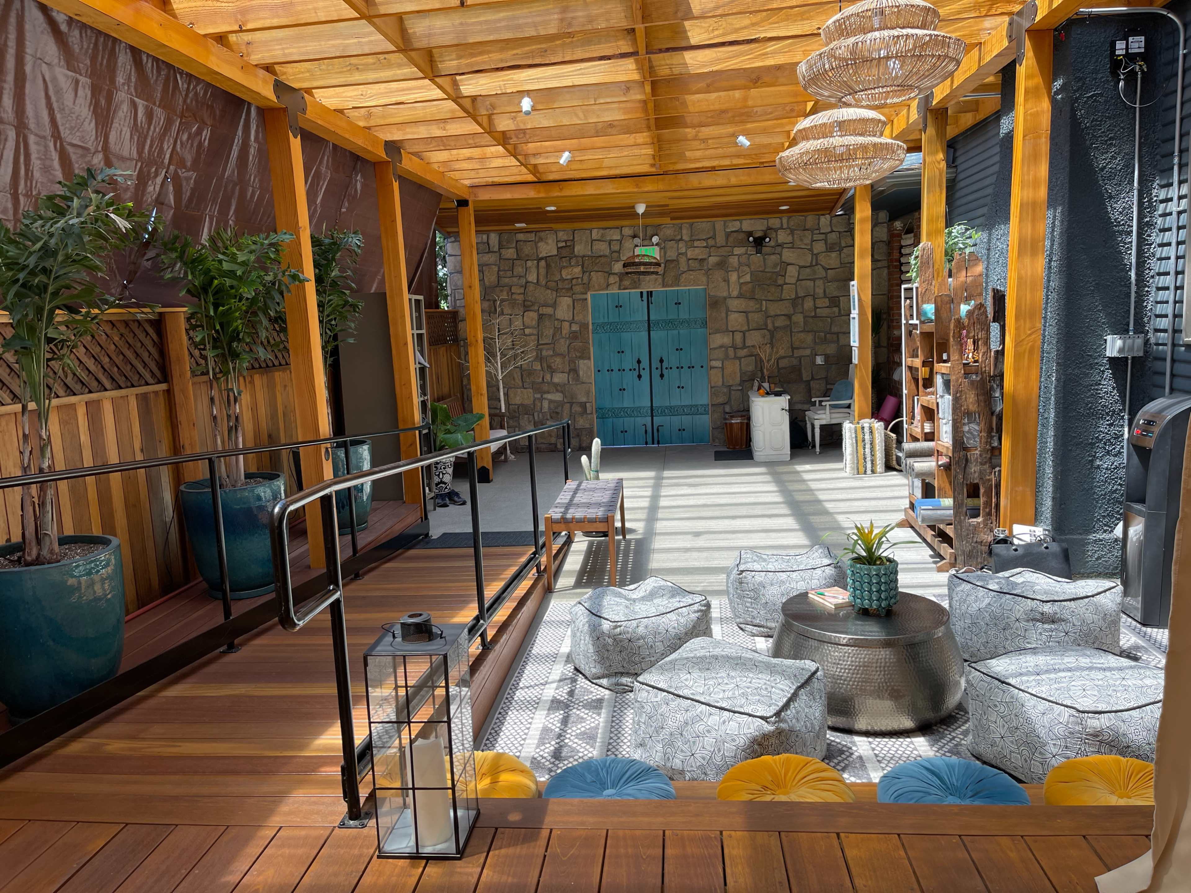 The image shows a well-lit indoor space featuring wooden beams, potted plants, a central seating area with poufs, and a stone wall with blue doors.