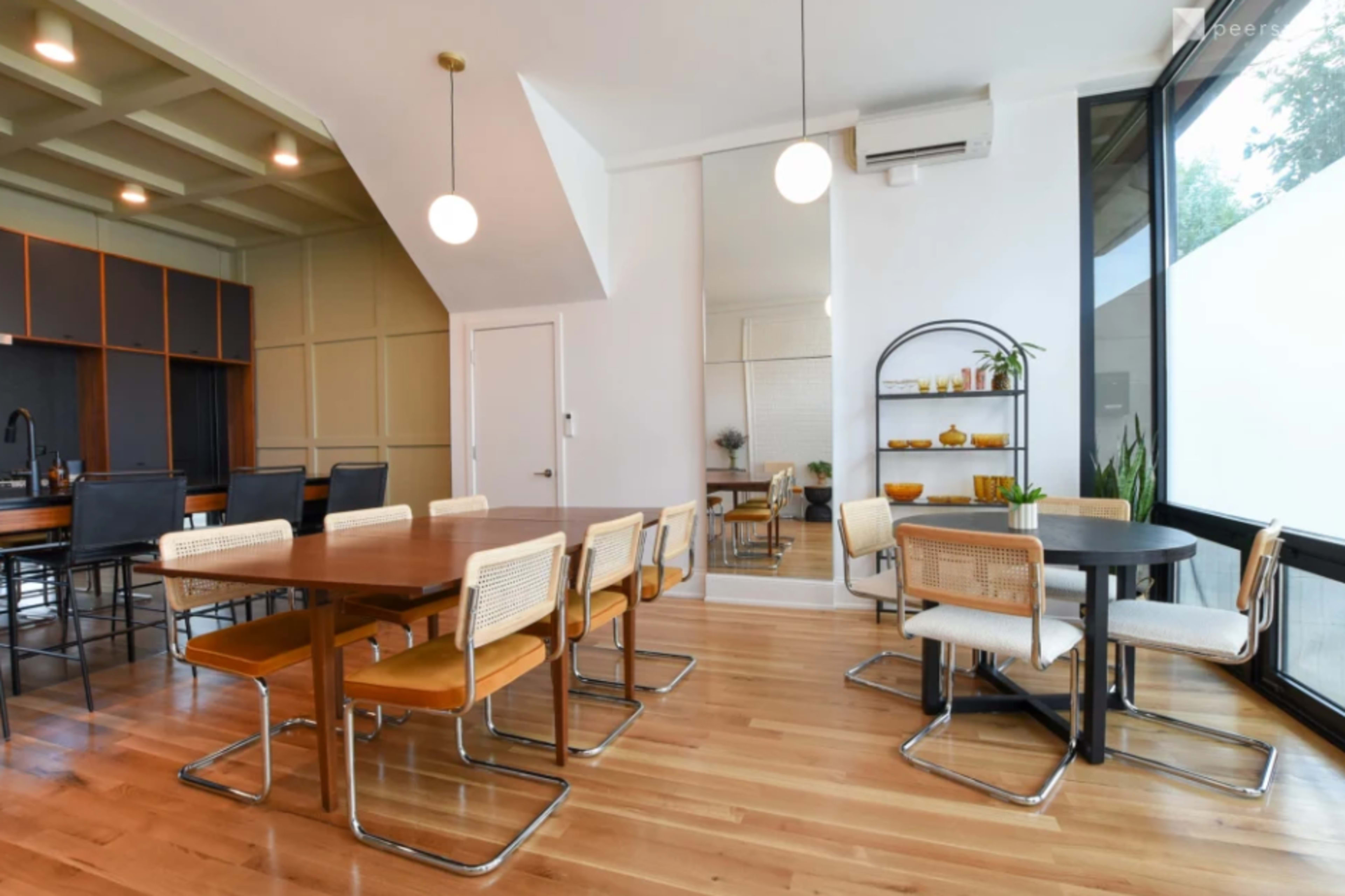 A modern dining area features a long wooden table with metal-framed chairs, a round table nearby, and large windows allowing natural light to fill the space.