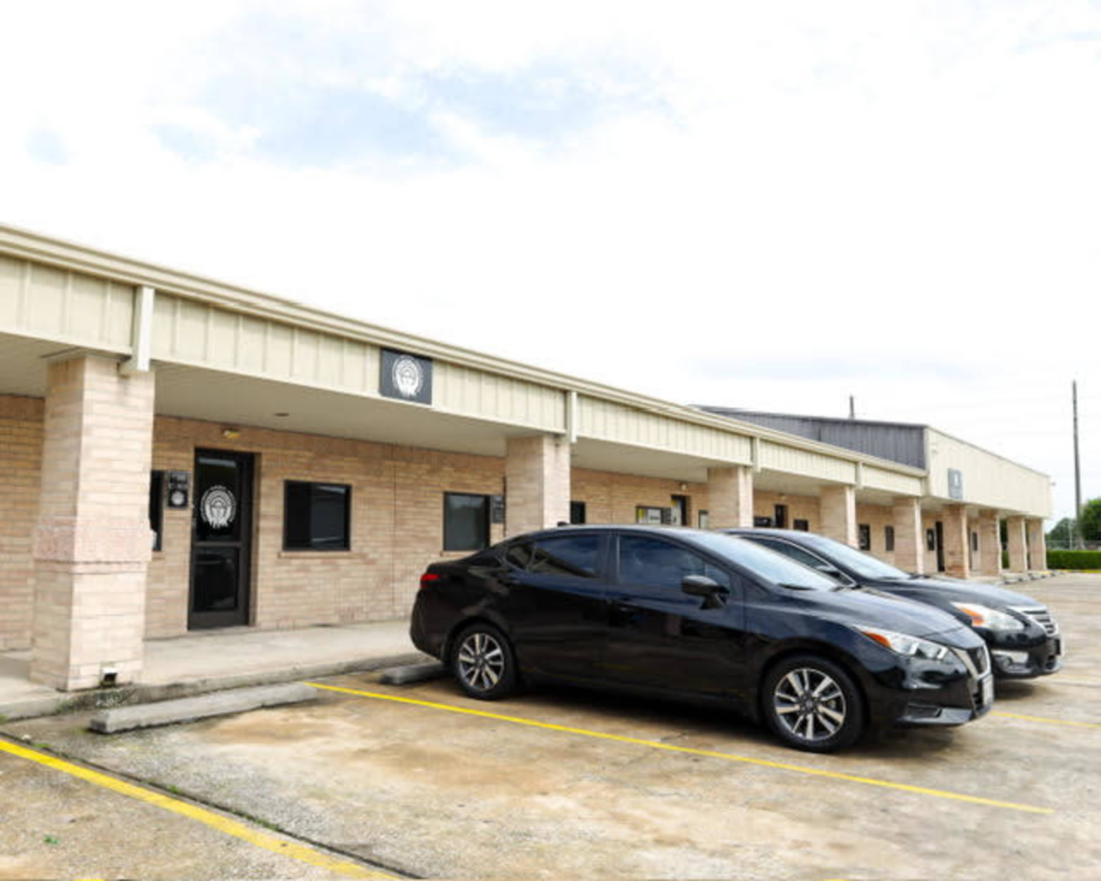 A row of commercial buildings with a parking lot containing two black cars.