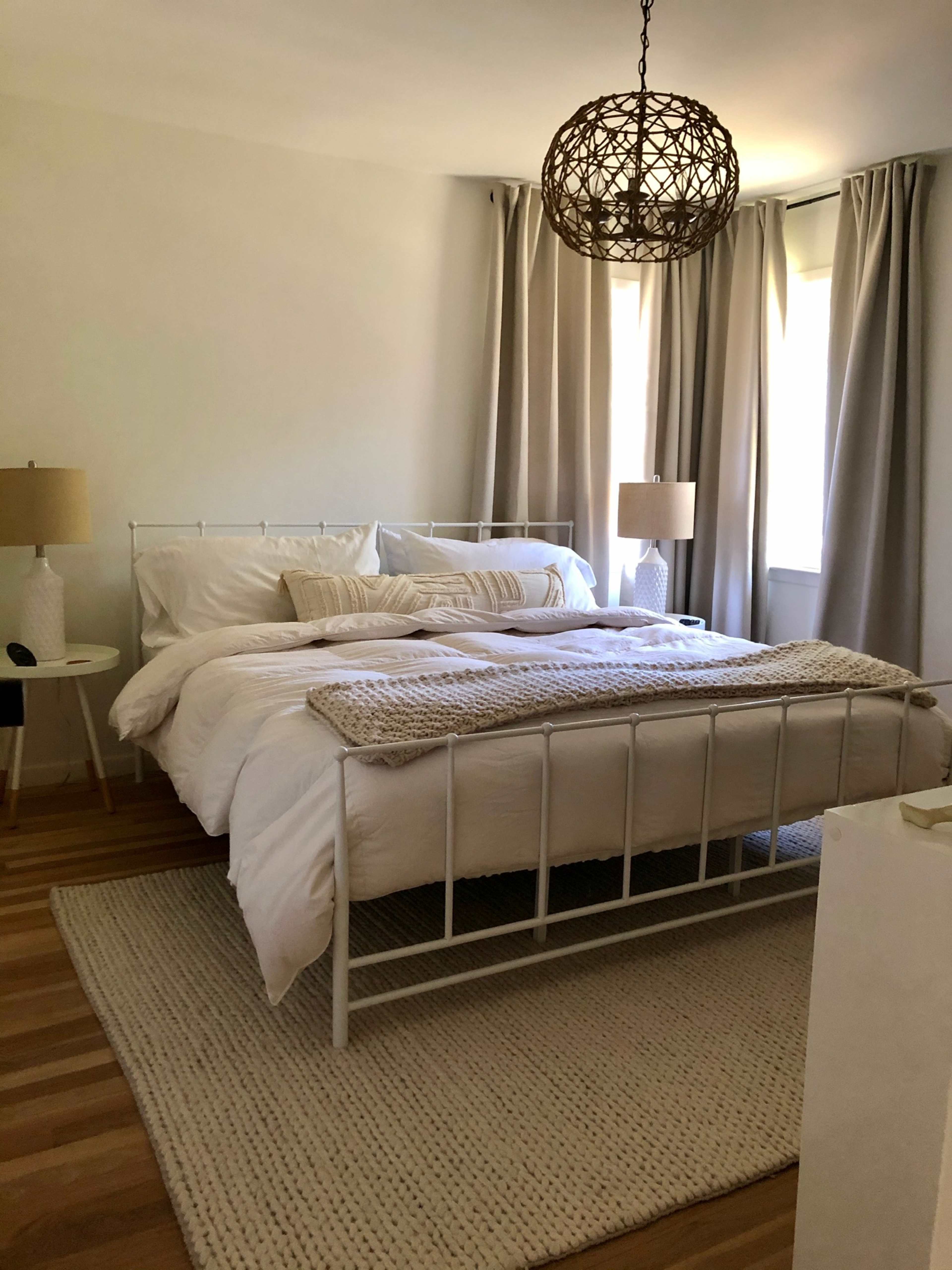 The image shows a softly lit bedroom featuring a white metal bed with linens, two bedside lamps, large windows covered with curtains, and a woven area rug on wooden flooring.