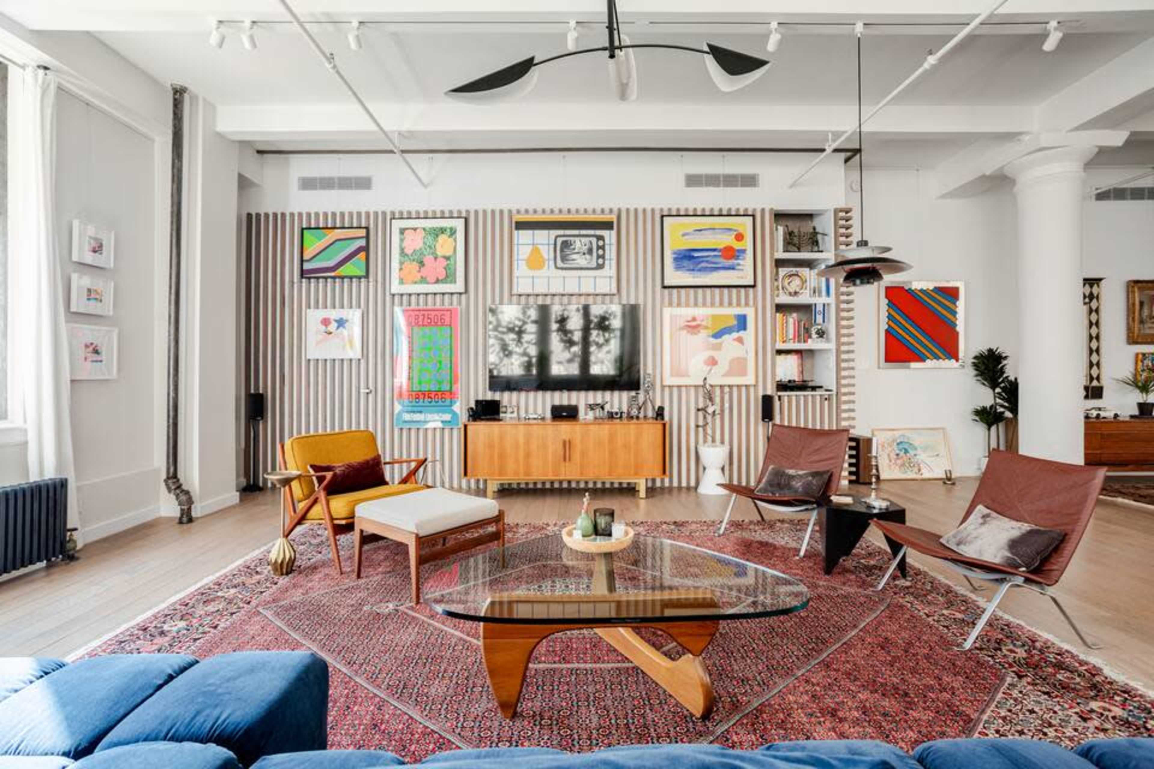 The image shows a modern living room decorated with colorful artwork, featuring a brown wooden media console, a glass coffee table, and various seating arrangements on a patterned rug.