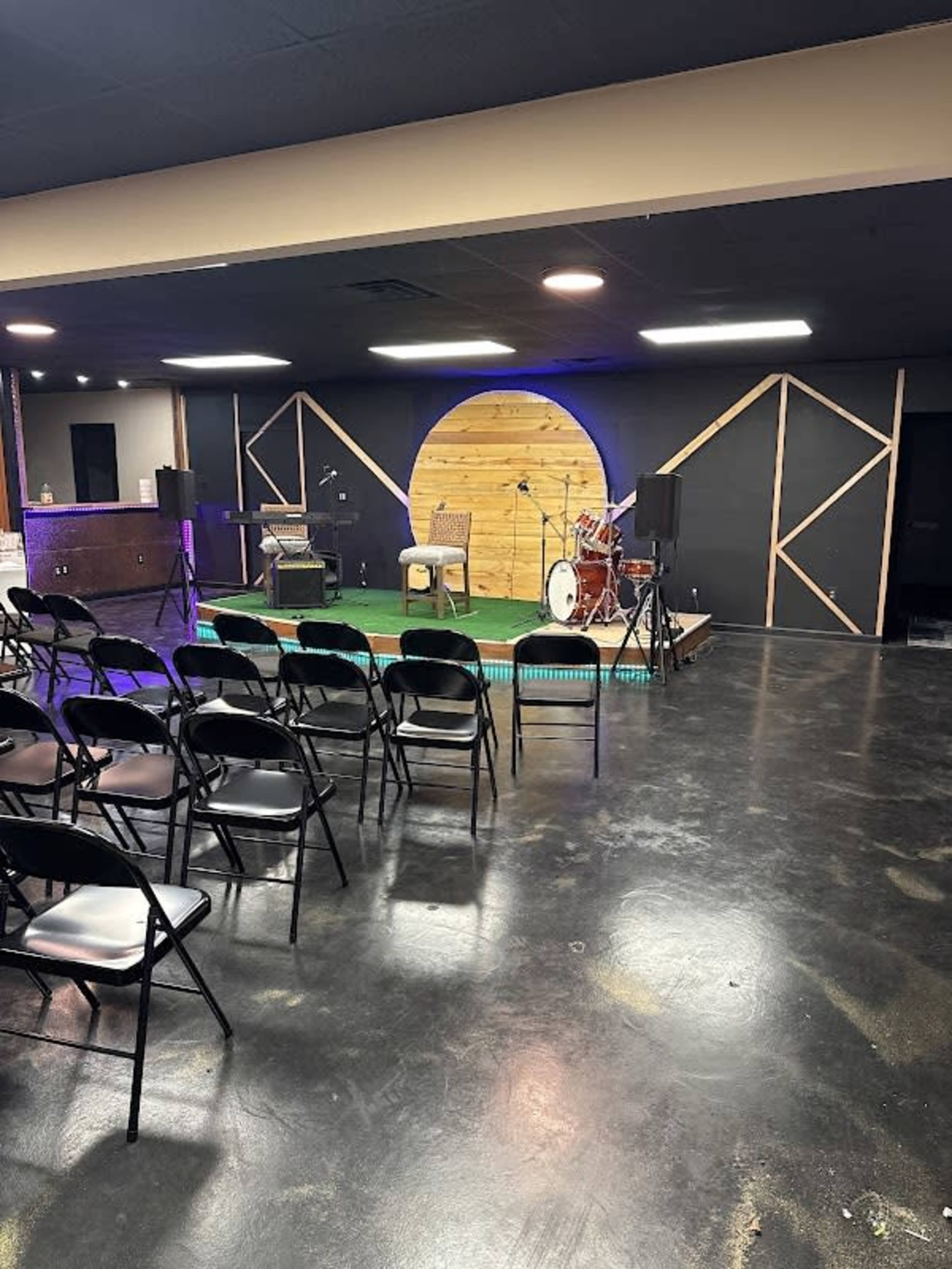 The image shows a small performance stage set up with a drum kit, a chair, and sound equipment in a dimly lit room arranged with black folding chairs facing the stage.