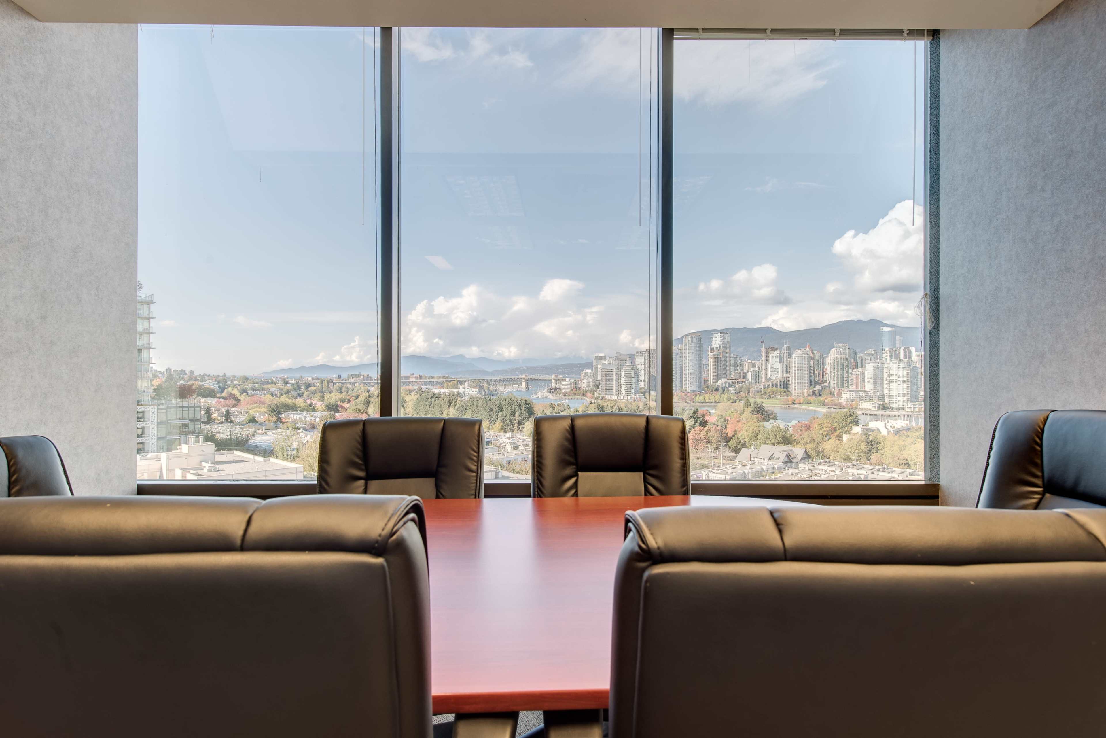 A conference room with a wooden table and black leather chairs, overlooking a city skyline and mountains through large windows.