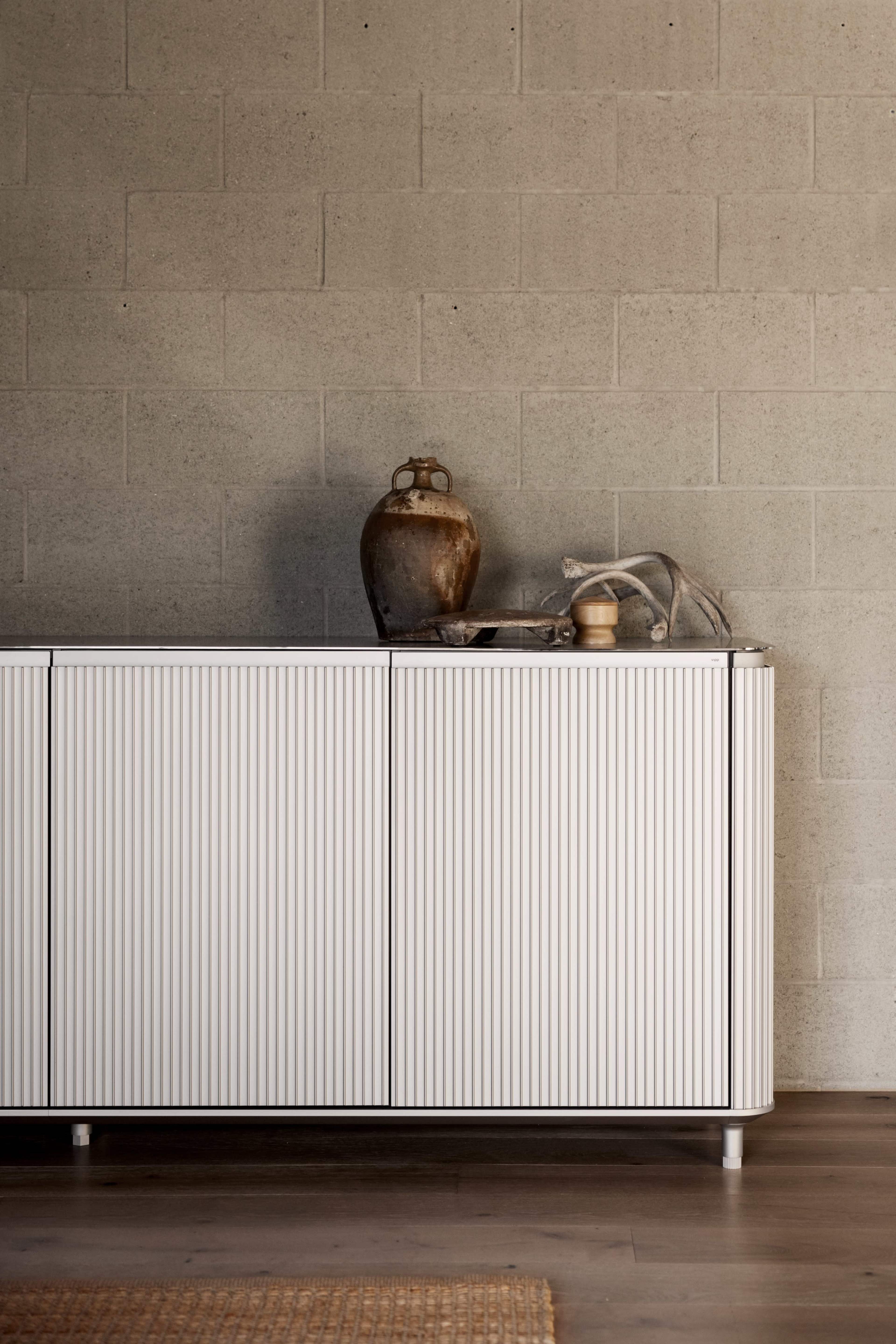 A modern, ribbed white cabinet sits against a gray, concrete block wall, with decorative pottery and a tray placed on top.