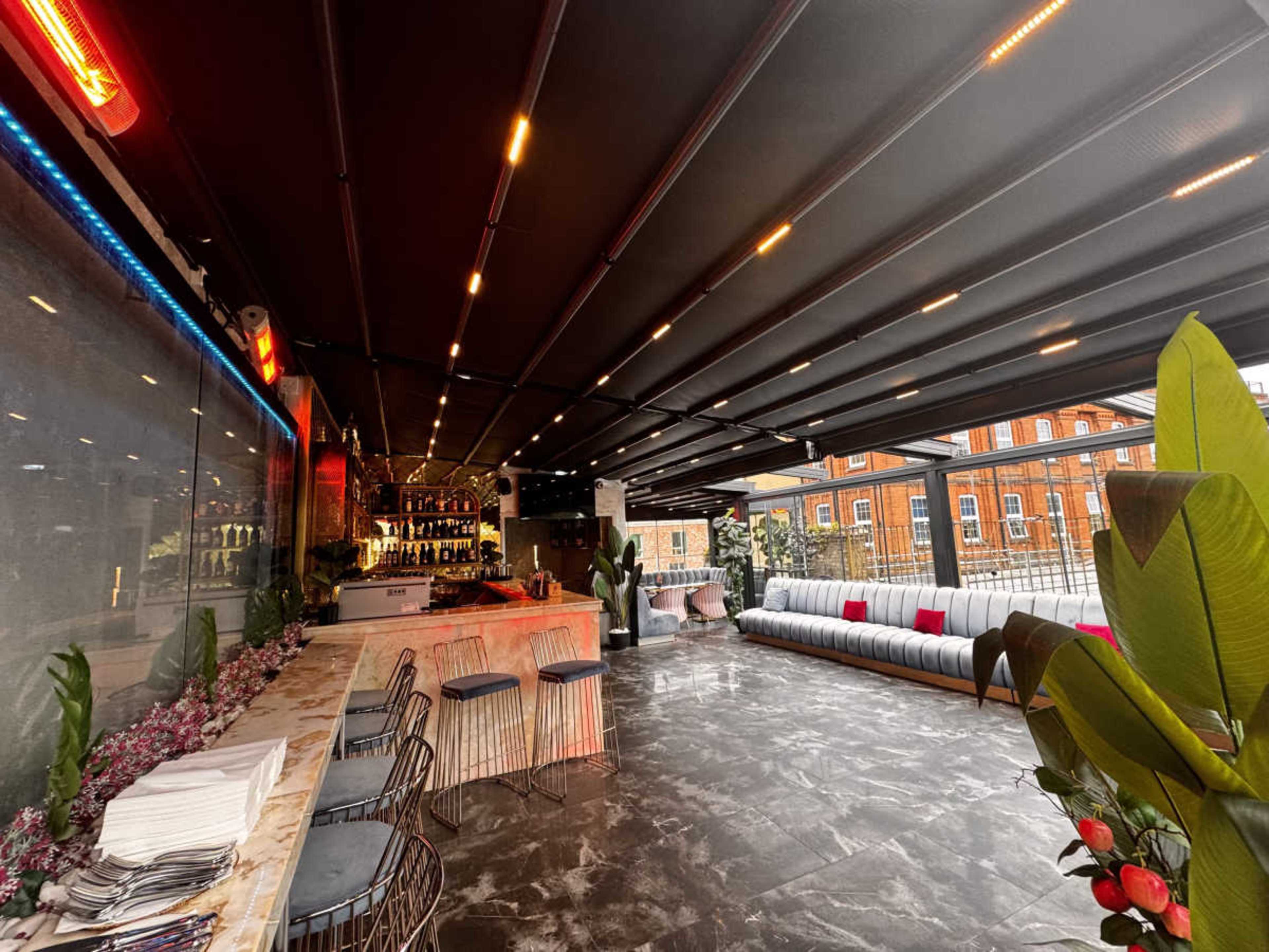 The image shows a modern indoor lounge area with a bar, sleek seating, and large windows providing a view of a red brick building.