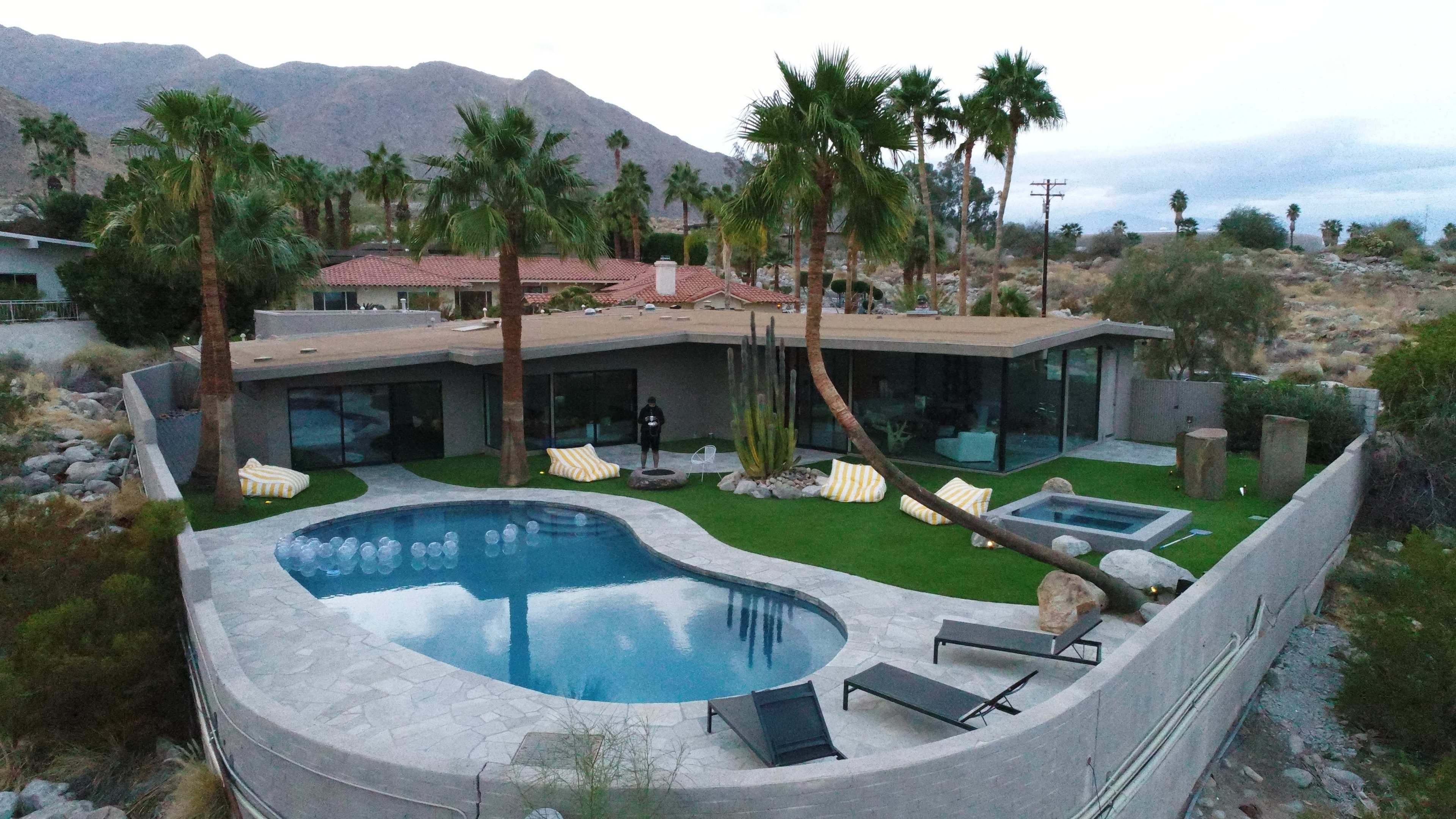 A modern house with large windows overlooks a curved swimming pool and surrounding palm trees in a desert landscape.