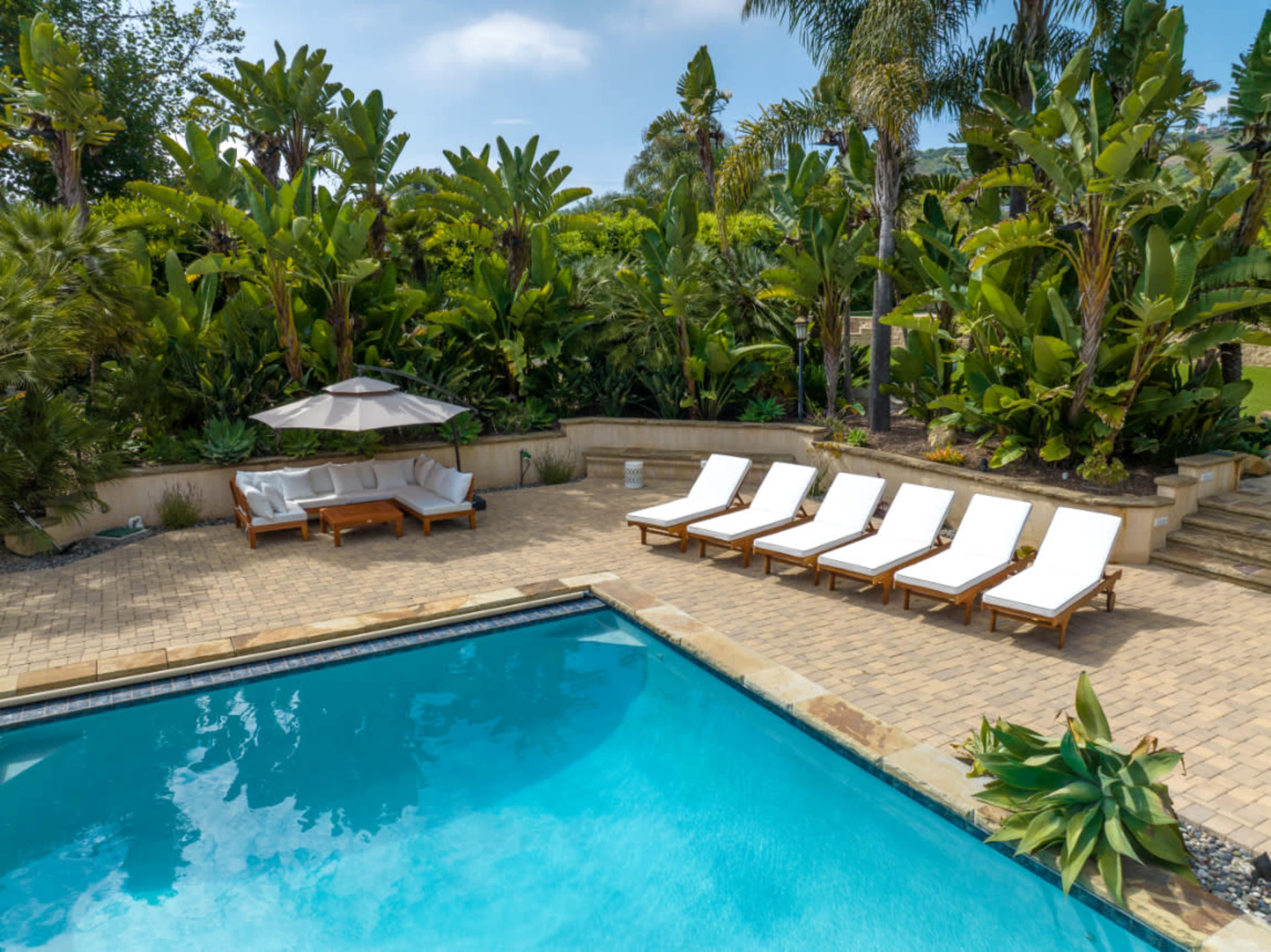 A swimming pool surrounded by lounge chairs and tropical plants is located in a landscaped outdoor area.