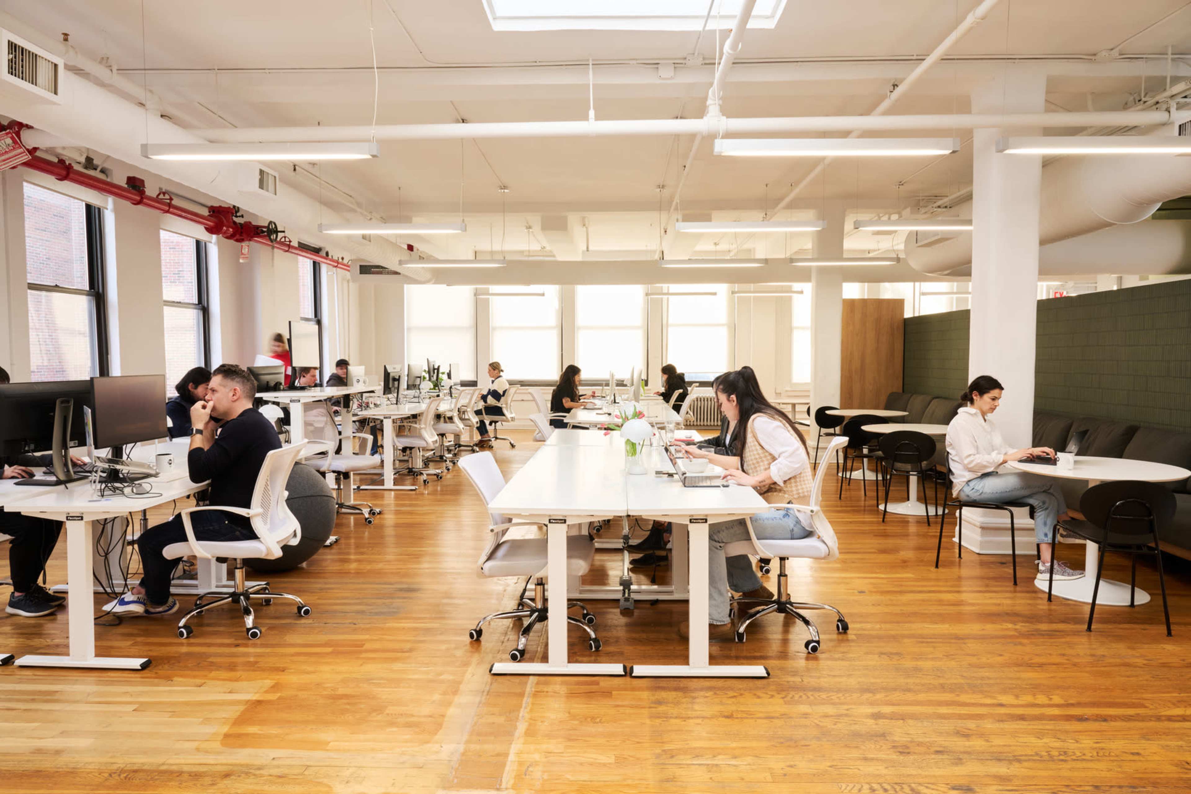 A modern office space features multiple workstations with people engaged in various tasks, surrounded by large windows and wooden flooring.