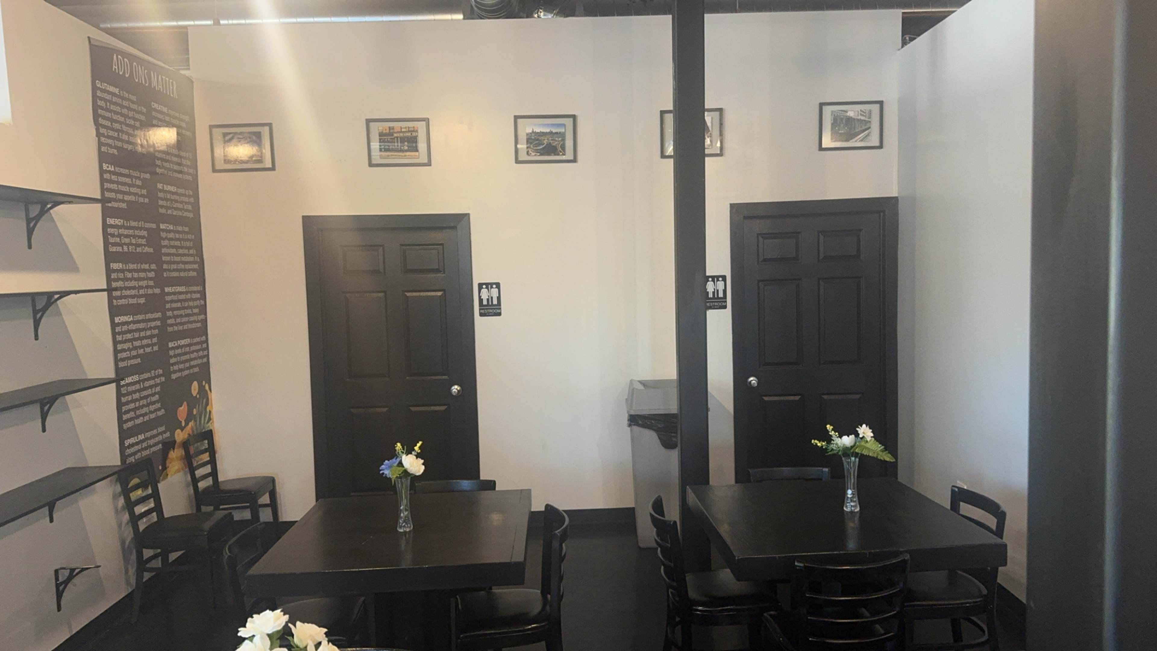 The image shows a restaurant interior with two tables, black chairs, and doors leading to restrooms on opposite walls, adorned with framed pictures.