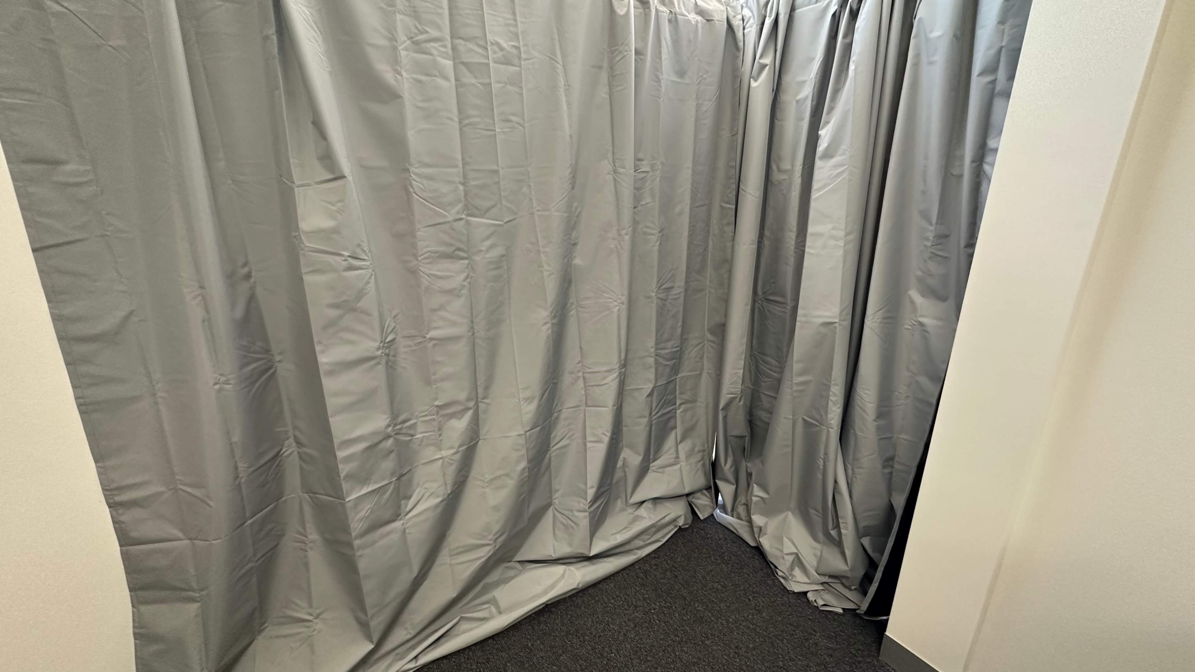 A corner of a room covered with gray curtains, partially draping over a dark carpeted floor.
