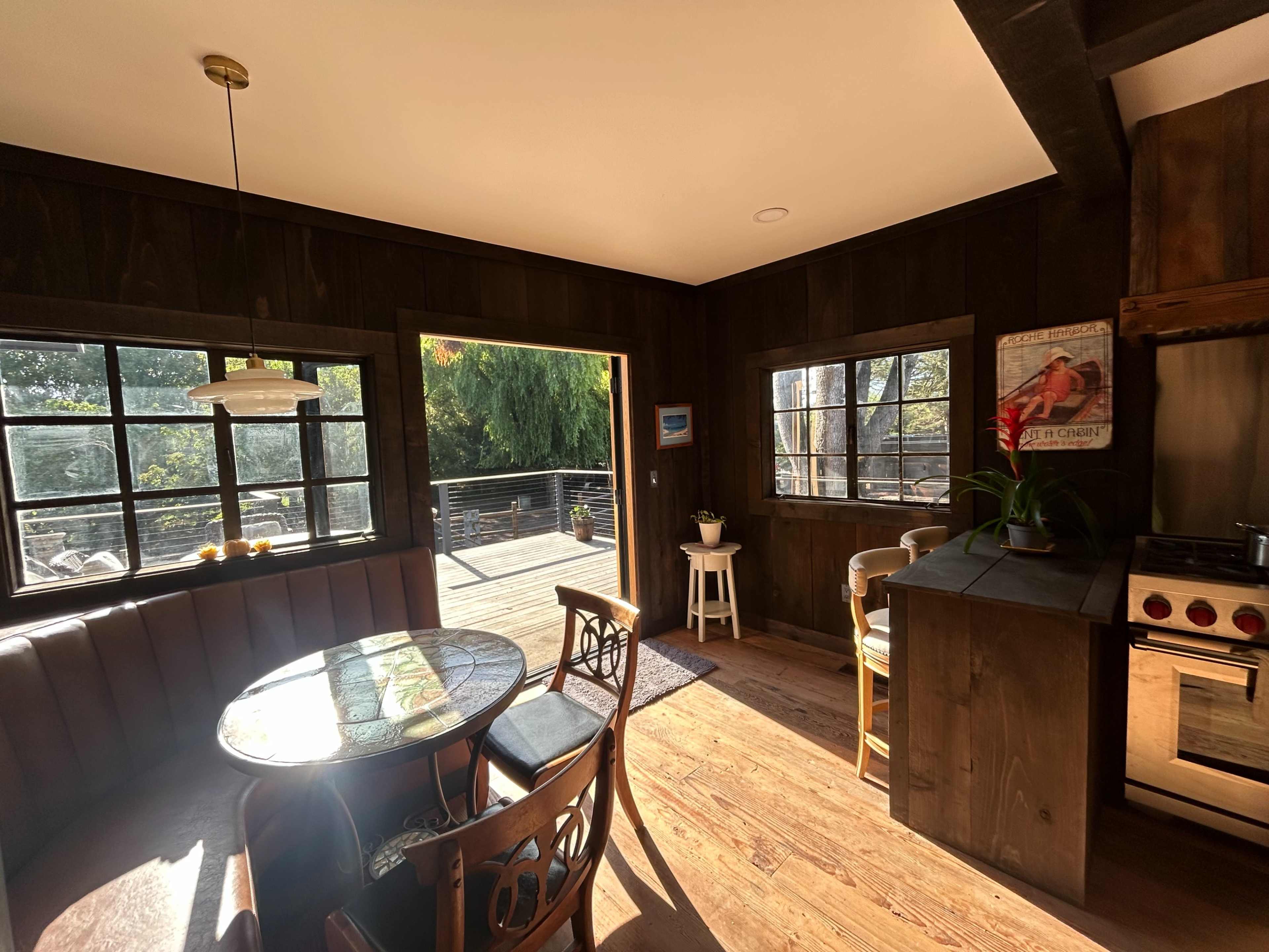 The image shows a sunlit kitchen with wooden walls, a round table with chairs, and an open doorway leading to a deck.
