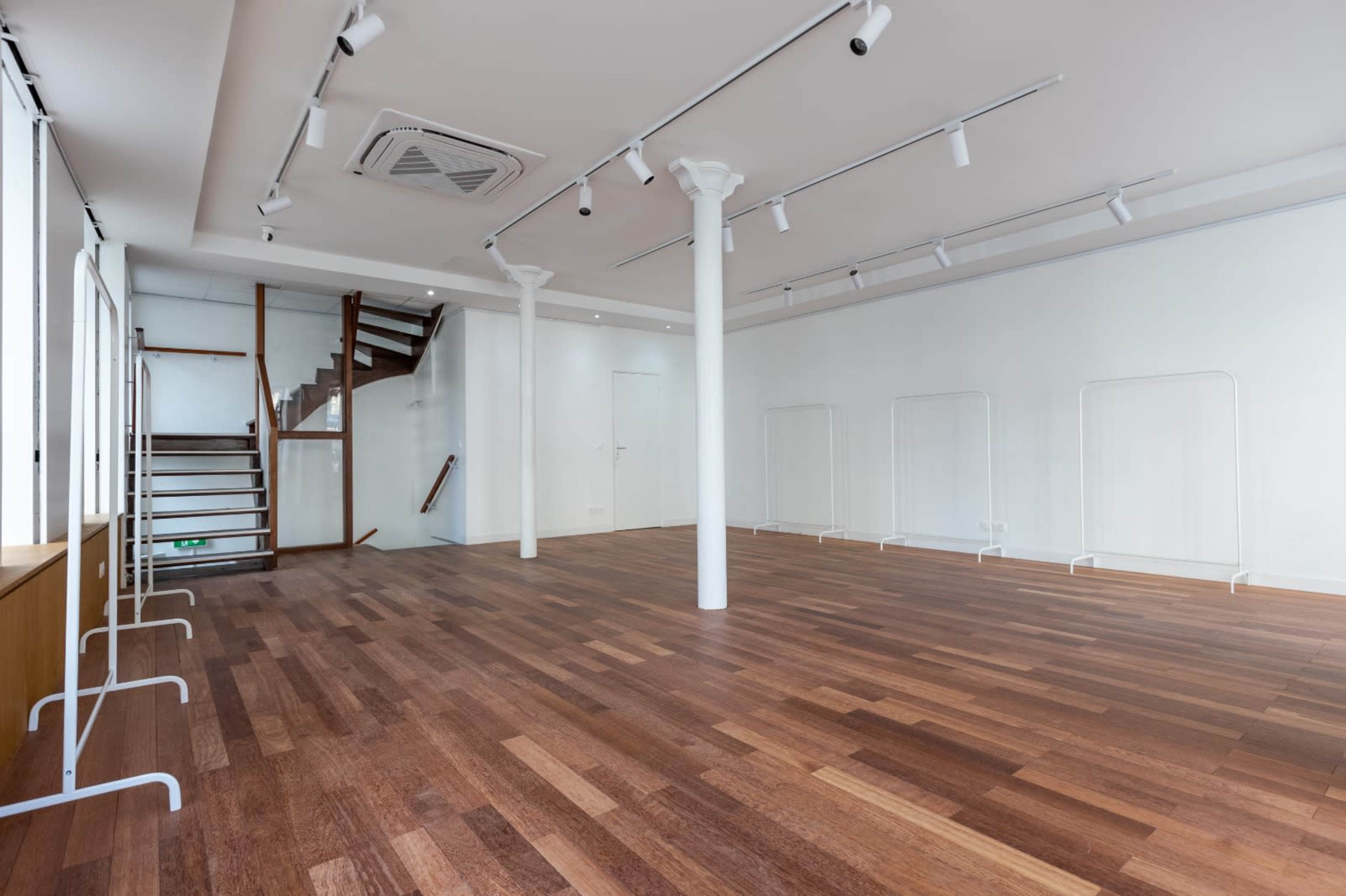 The image shows an empty interior space with wooden flooring, white walls, and a staircase in the background.