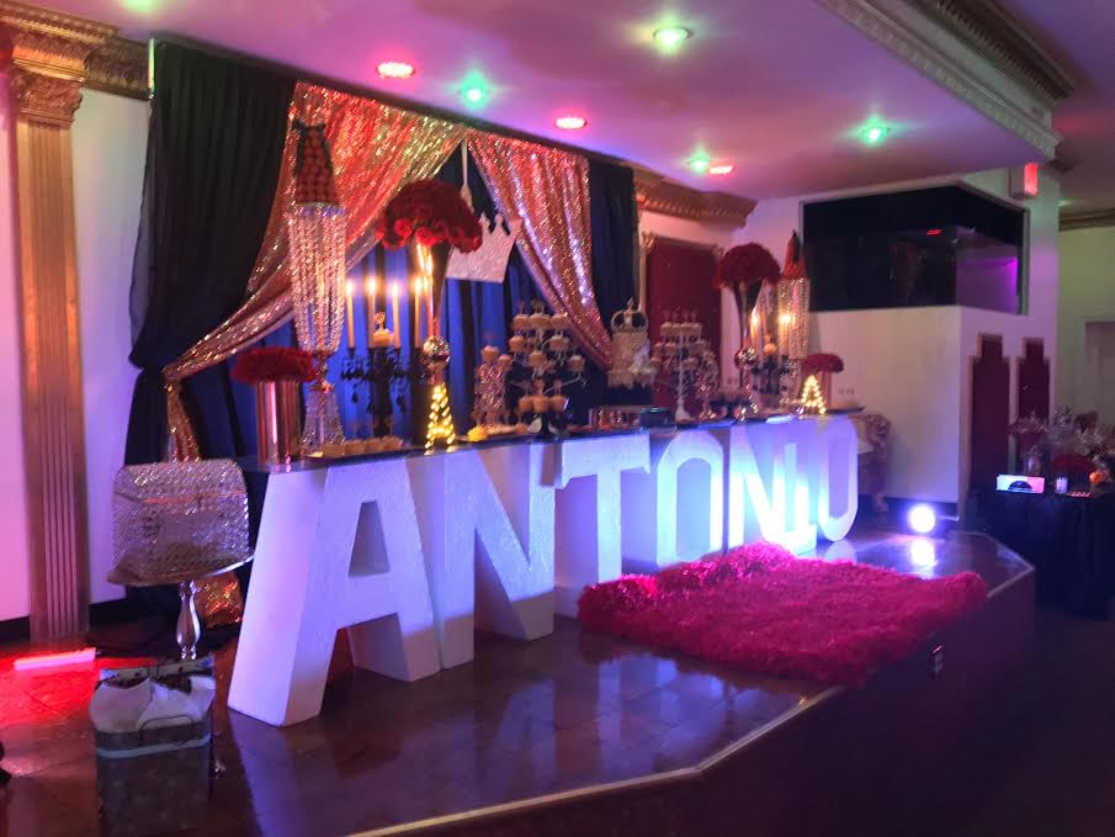 The scene features a decorative event display with a large sign spelling "ANTONIU," surrounded by elegant decorations, tall candles, and red floral arrangements.