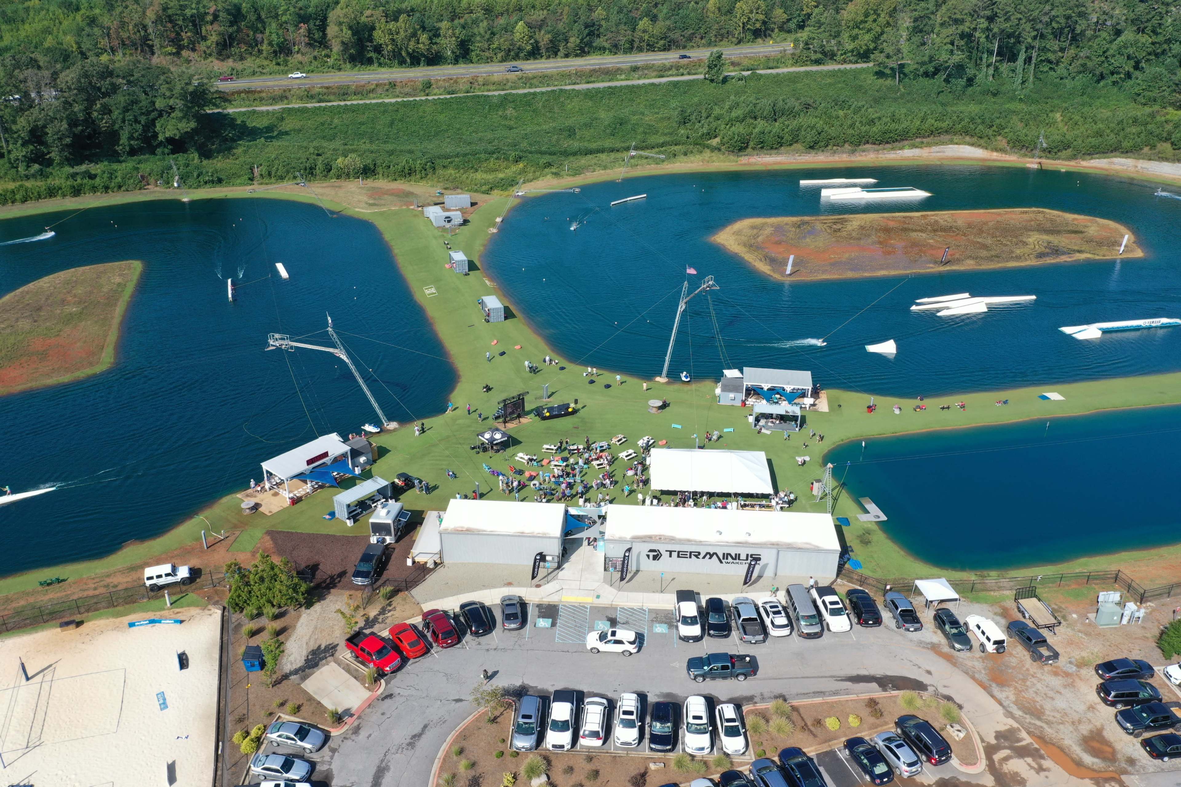 An aerial view shows a cable wakeboarding facility with water for skiing, surrounding grassy areas, and a parking lot filled with cars.