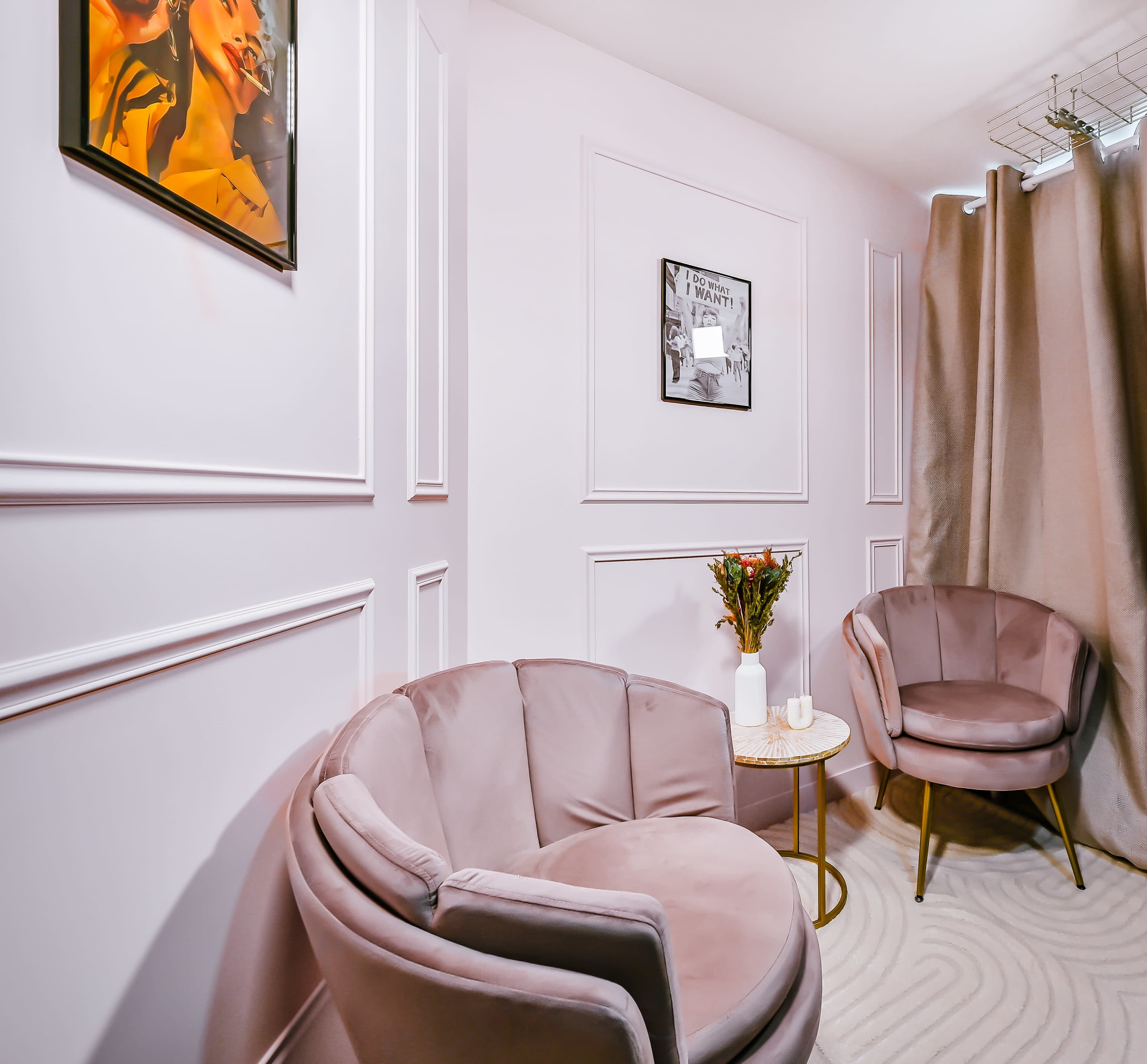 The image shows a softly lit room featuring two pink velvet armchairs, a small round table, and framed artwork on the walls, with a curtain draped in one corner.