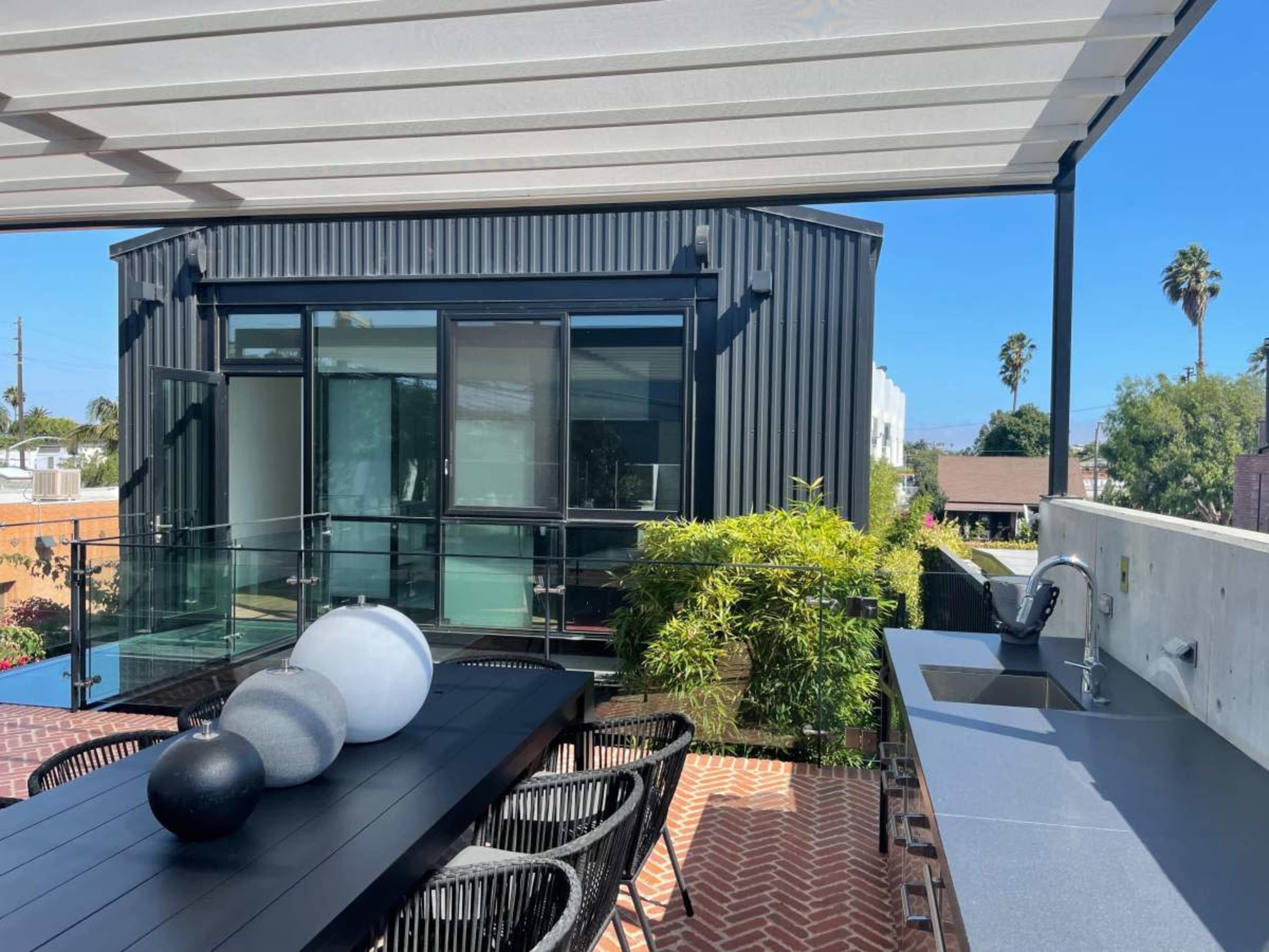 A modern outdoor dining area with a black table, three decorative spheres, and a sleek kitchen counter, overlooking a building with large windows and tropical greenery.