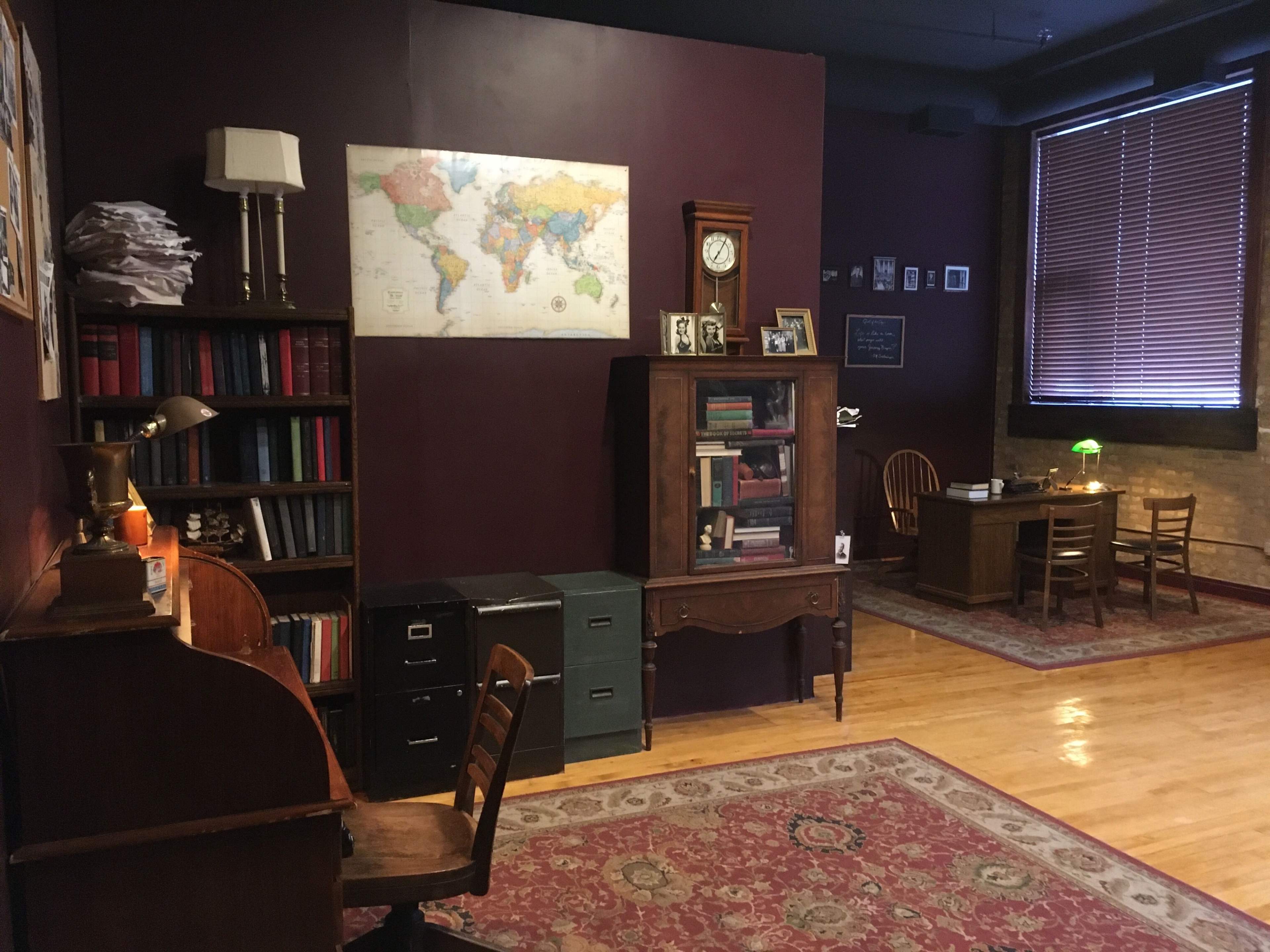 The image shows a cozy room featuring a map of the world on the wall, a wooden desk with a chair, a bookshelf filled with books, a lamp on a side table, and a small table with two chairs.