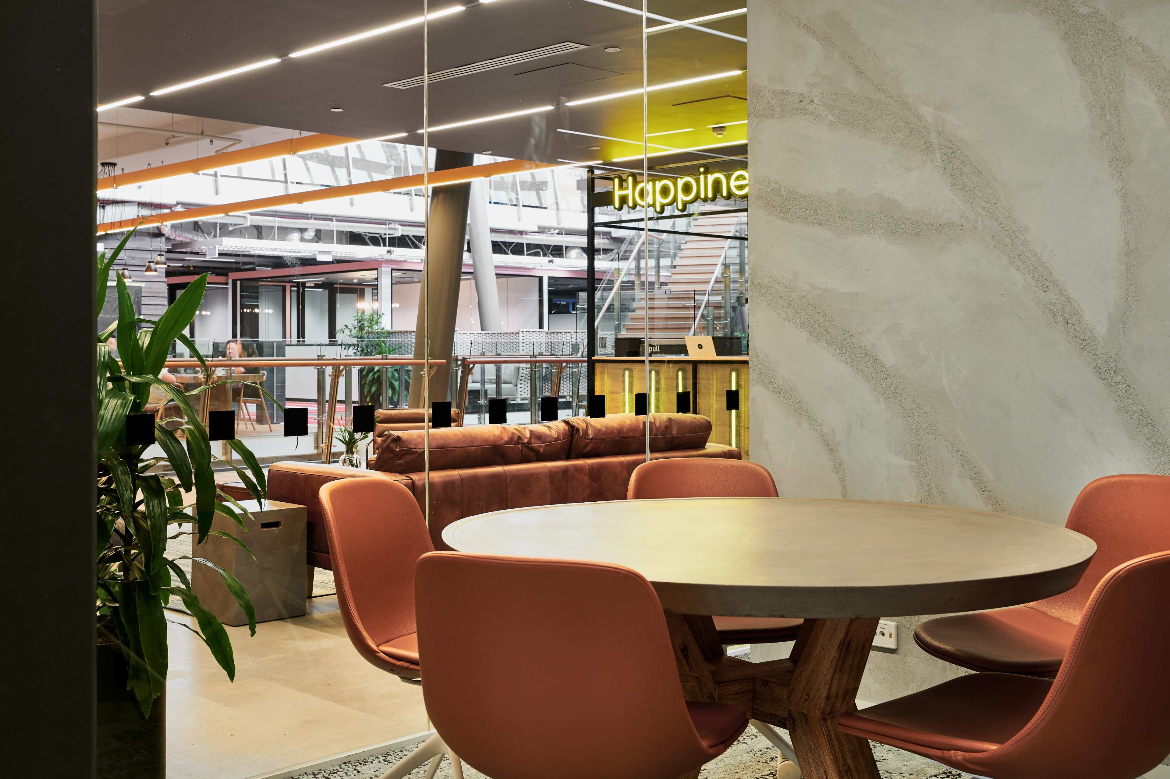 The image shows a modern office space featuring a round wooden table surrounded by orange chairs, with a cozy seating area in the background and a neon sign that says "Happiness."