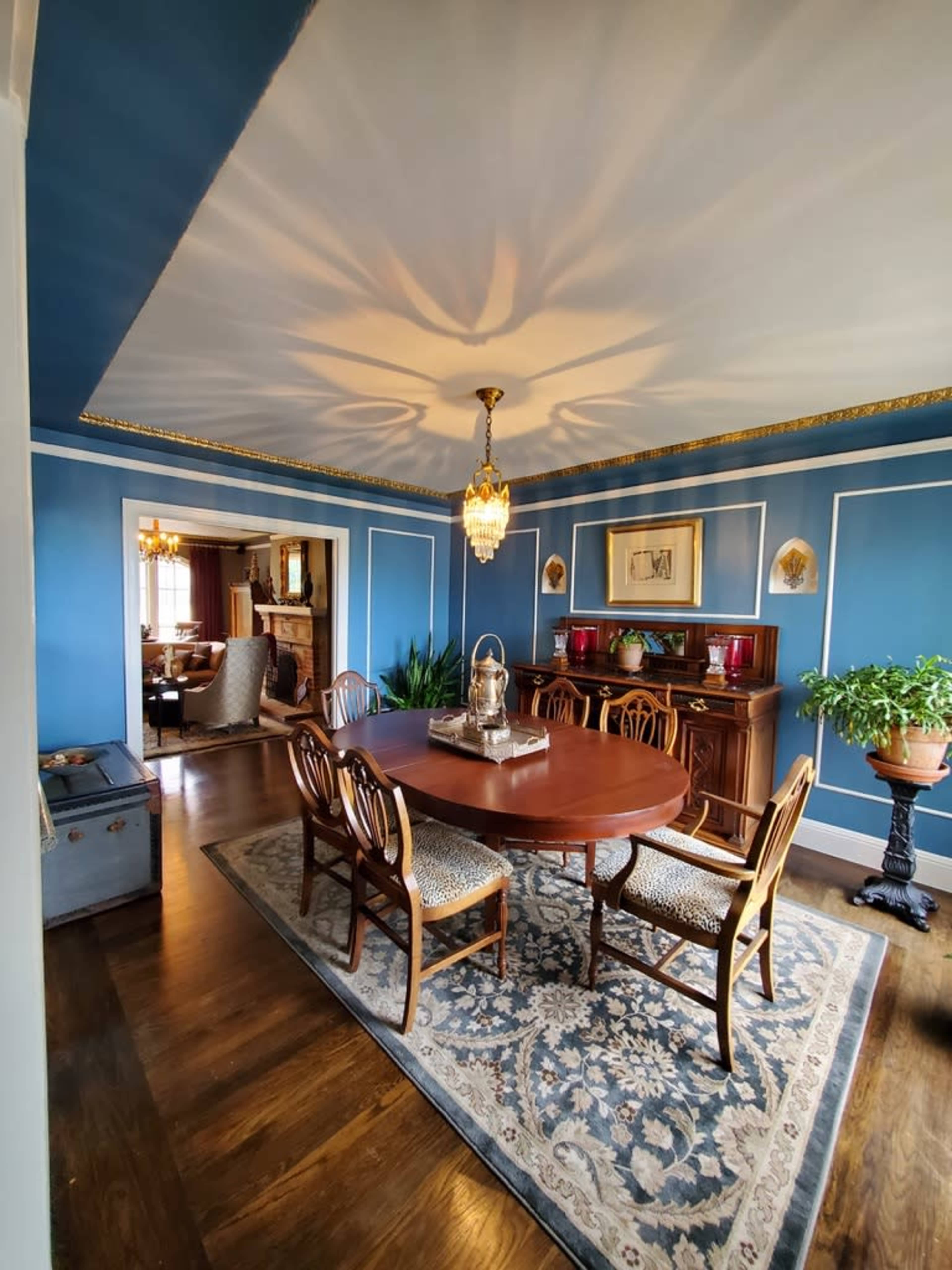 A dining room with a wooden table surrounded by chairs, blue walls, a chandelier, and a decorative rug.