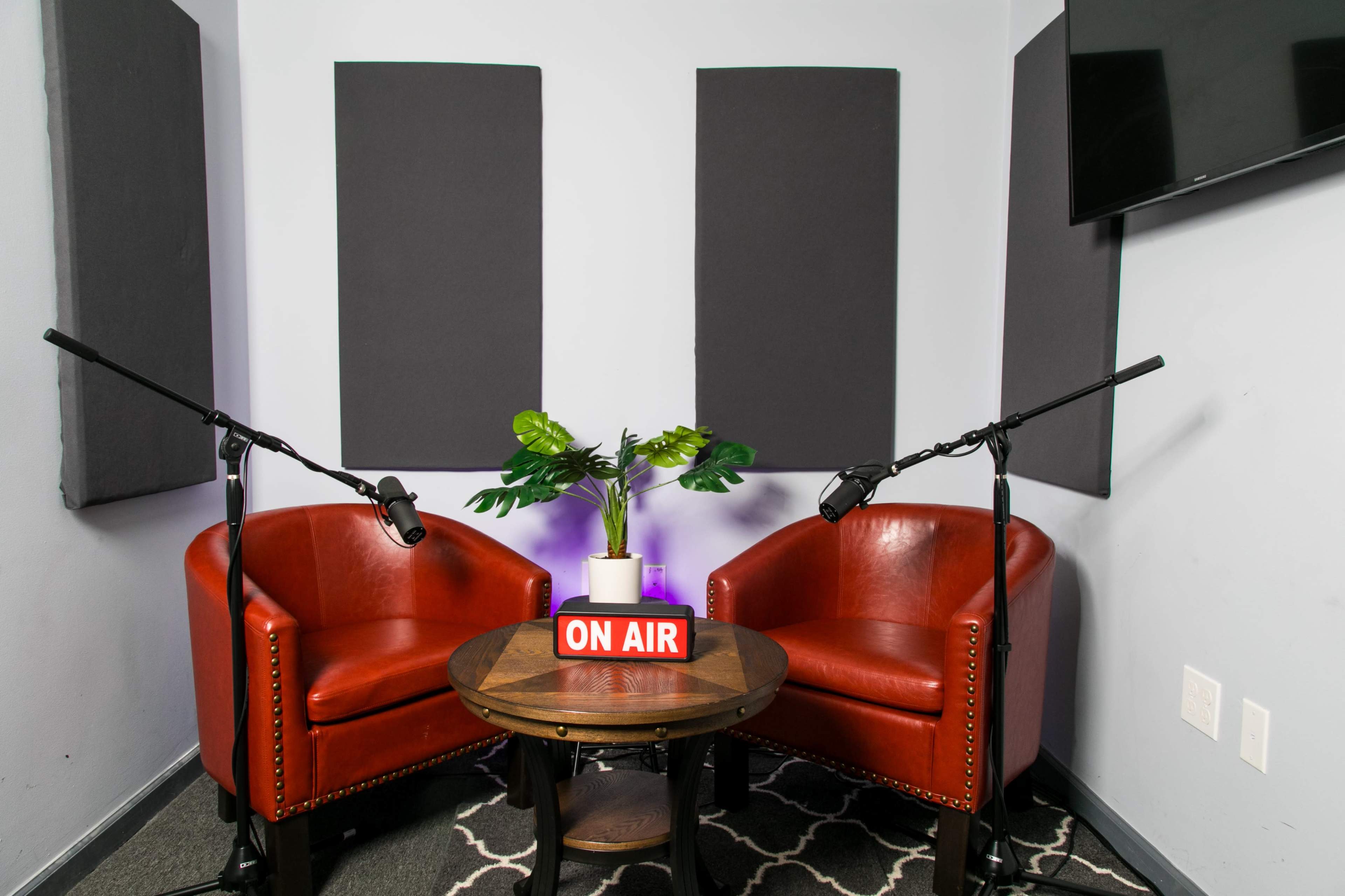 A small recording studio setup with two red chairs facing each other, a round table with a "ON AIR" sign, and microphones positioned on either side.