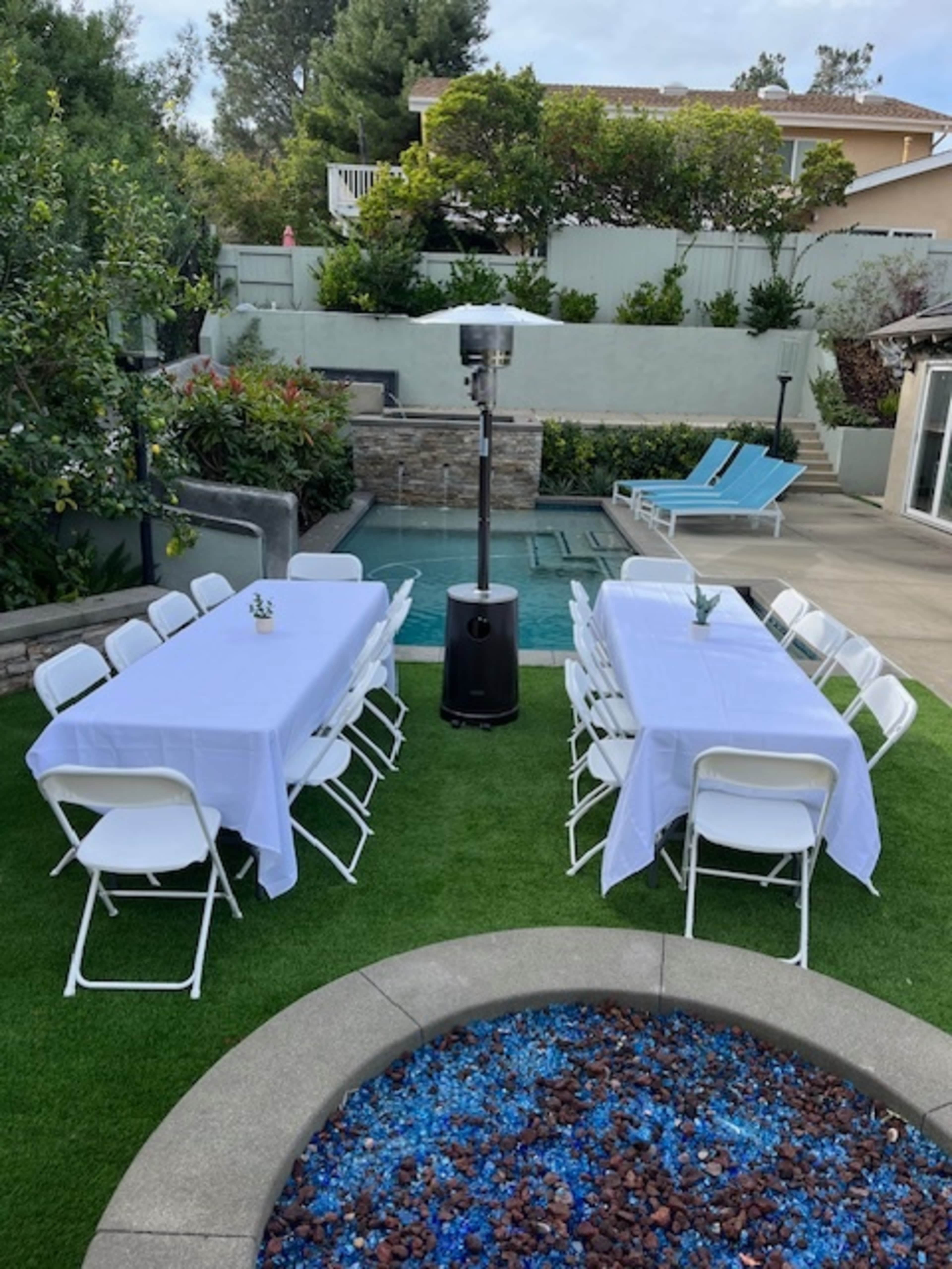 An outdoor gathering area features two tables set with white tablecloths, surrounded by greenery and a pool in the background.
