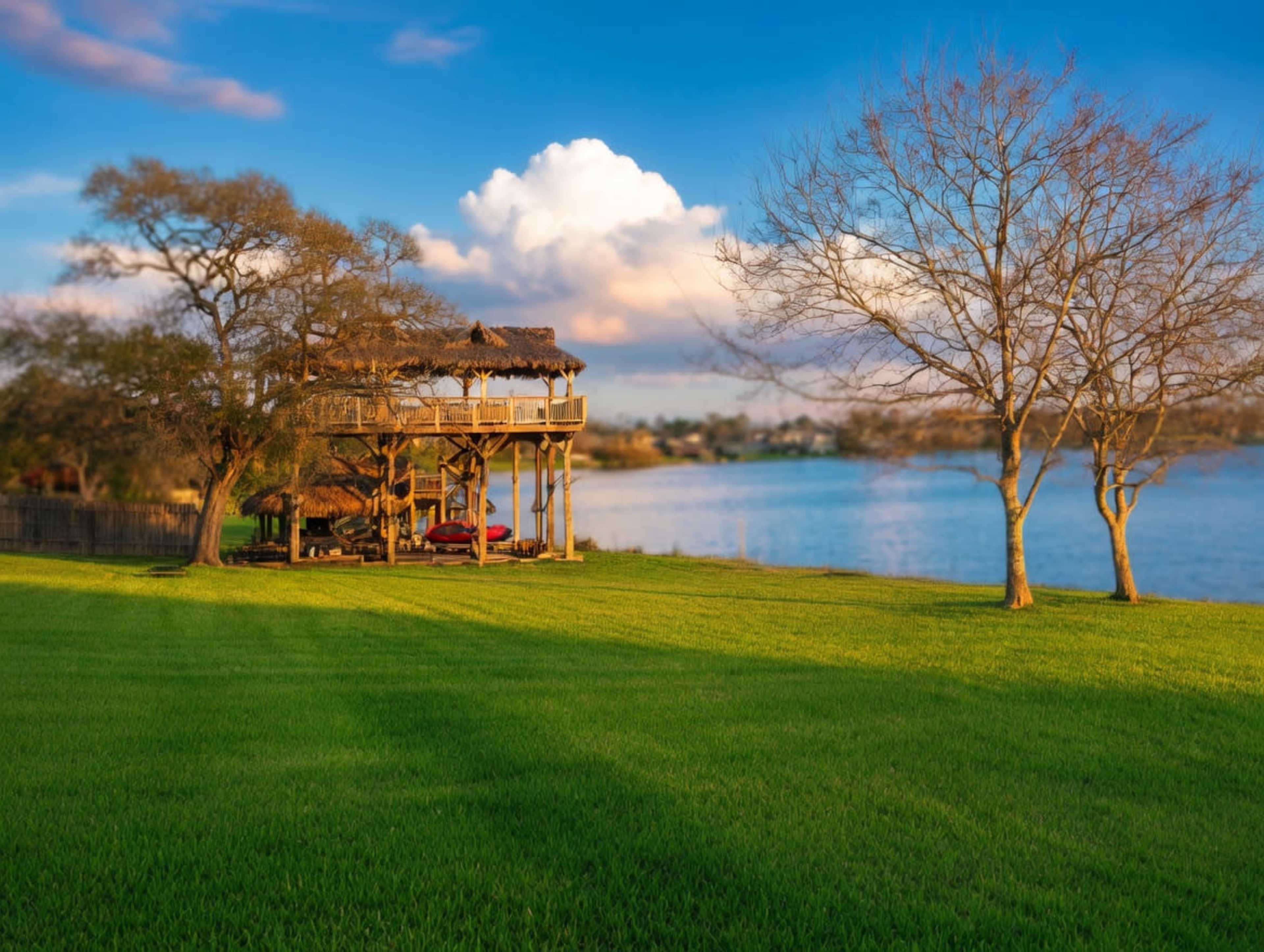 A wooden structure with a thatched roof overlooks a calm body of water, surrounded by green grass and a few trees under a blue sky with clouds.