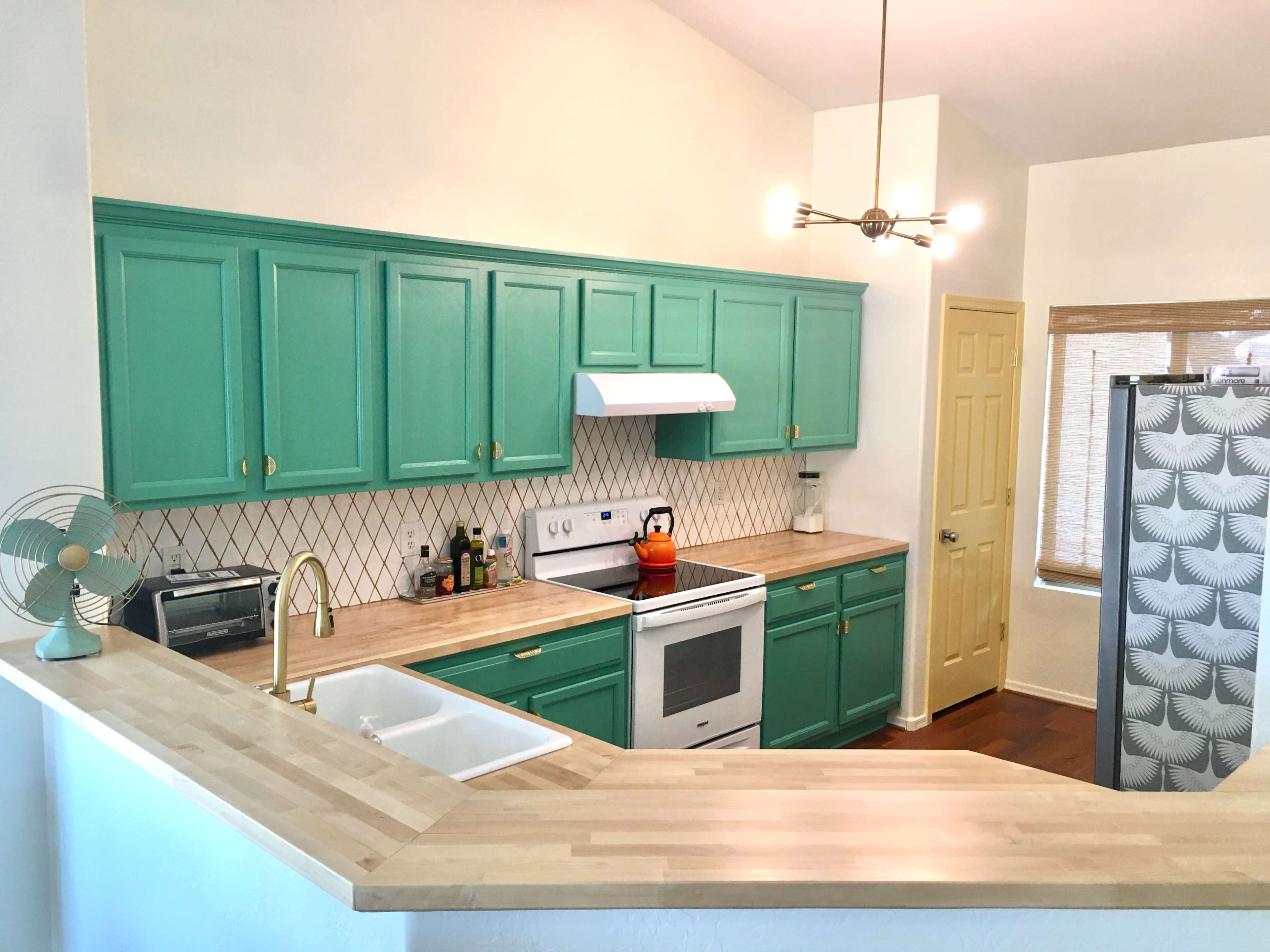 A modern kitchen with teal cabinets, a white stove and sink, and a stainless steel refrigerator, illuminated by overhead lighting.
