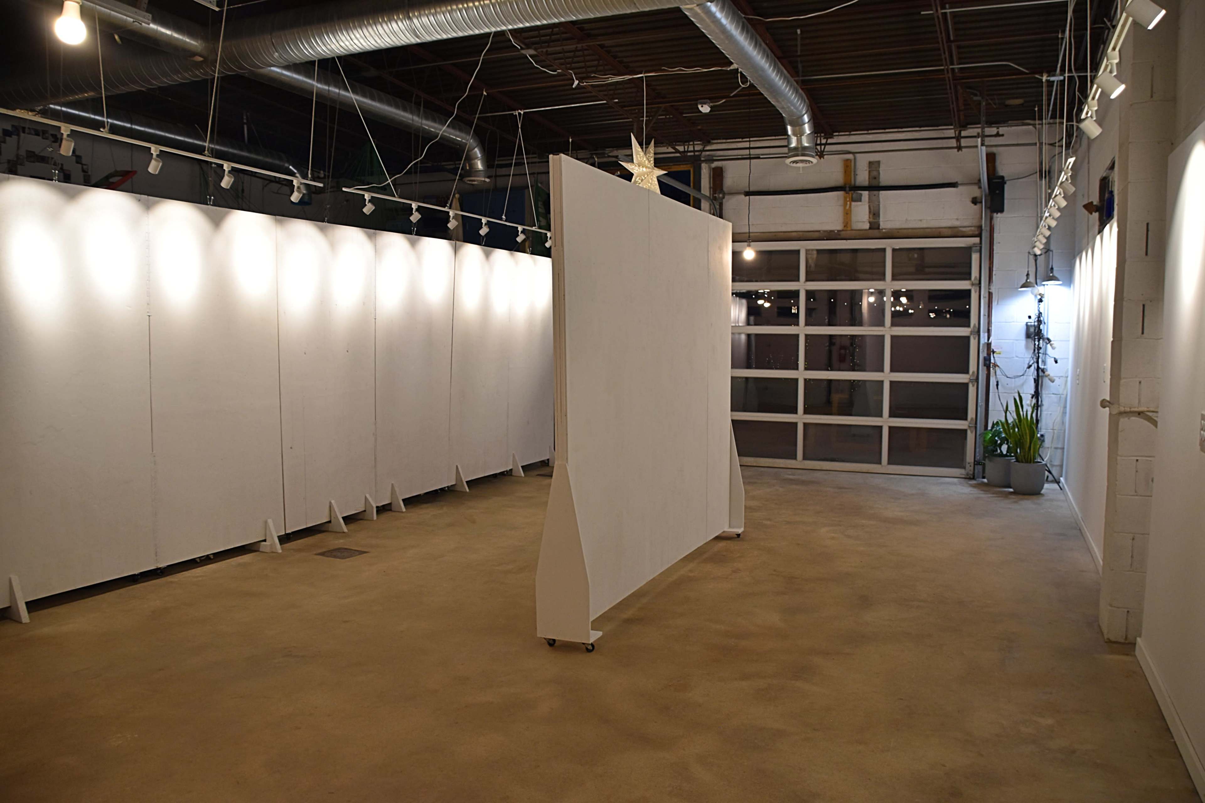 A spacious art gallery with white walls, a movable partition in the center, and a large garage door at one end.