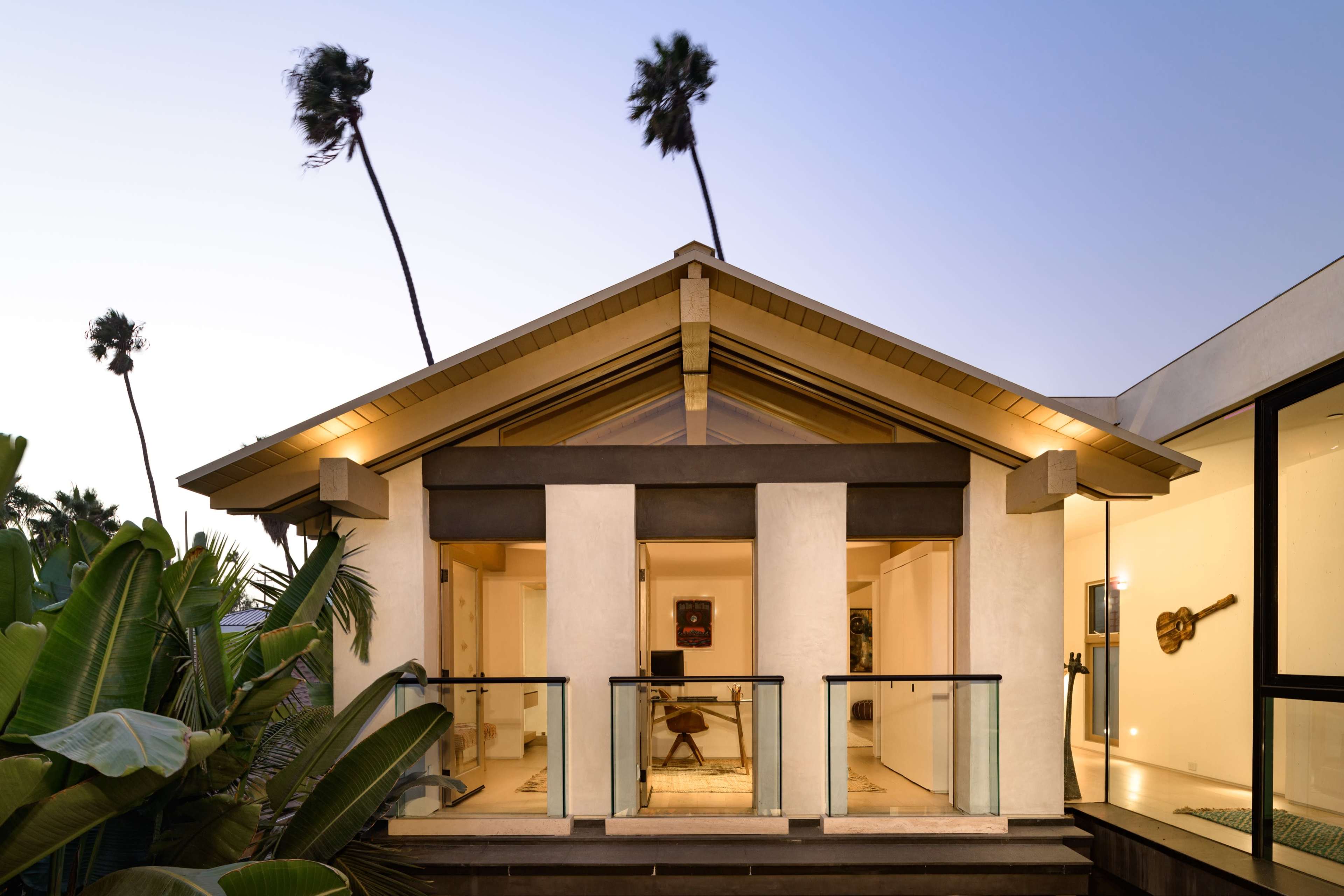 A modern house features a distinctive architectural style, with a sloped roof and large glass doors opening onto a balcony surrounded by palm trees.