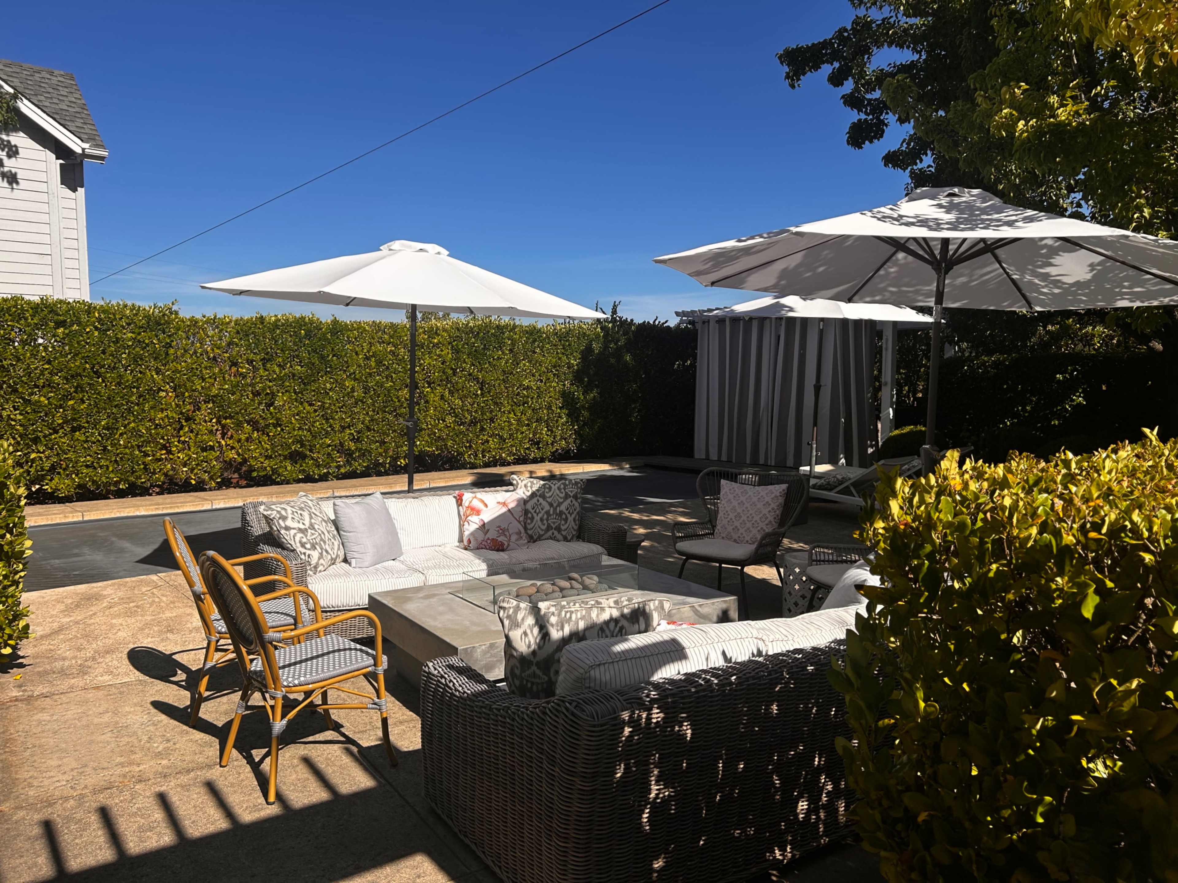 The image shows a sunlit outdoor seating area with white umbrellas, surrounded by hedges and a concrete patio, featuring various lounge chairs and a sectional sofa.