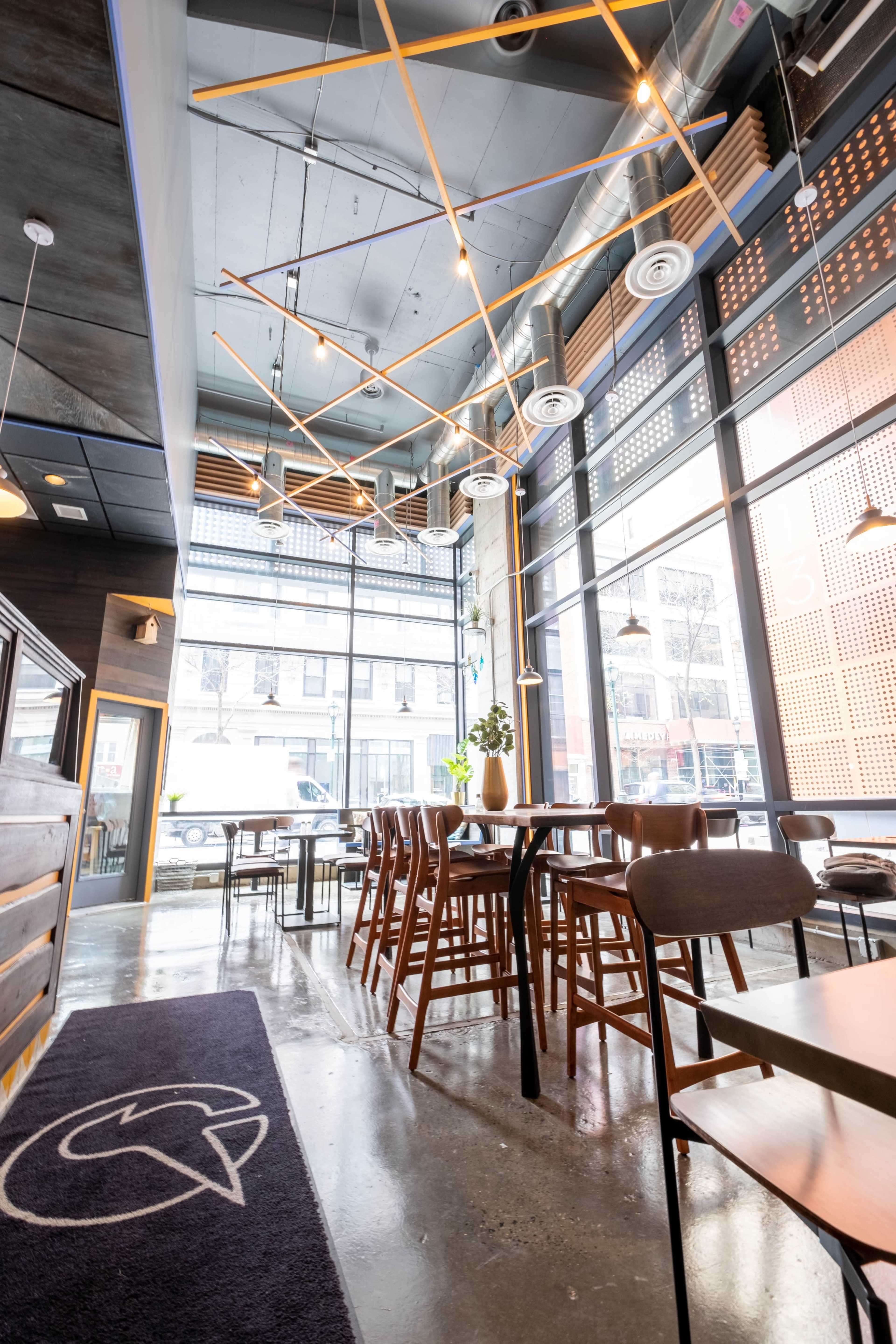 A modern café interior featuring wooden furniture, large windows, and decorative ceiling lights.